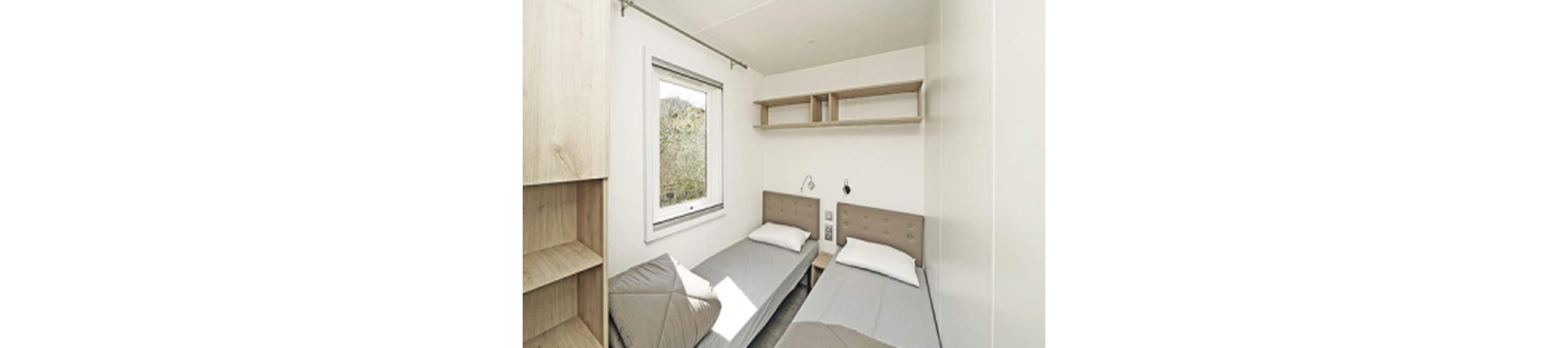 A compact bedroom features two single beds with neutral-colored headboards and simple white bedding. Natural light enters through a window on the left, illuminating the room and the view of greenery outside. Built-in wooden shelving and a small wall-mounted reading light emphasize the minimalist, functional design. The overall palette is light and airy, with soft beige and white tones creating a calm atmosphere.