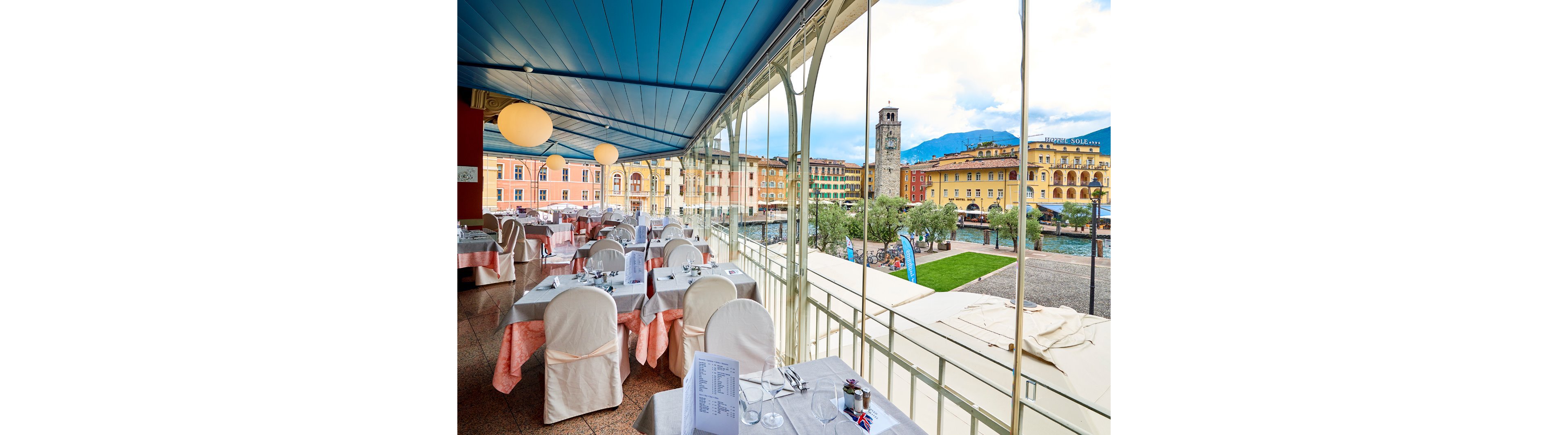 An elegant restaurant terrace overlooks a colorful waterfront town square. Tables are neatly set with white cloths, covered chairs, and glassware beneath a blue ceiling with round hanging lights. Large floor-to-ceiling windows frame historic buildings, a stone tower, and distant mountains under a partly cloudy sky. The scene feels bright and inviting, combining urban architecture with a relaxed dining atmosphere.