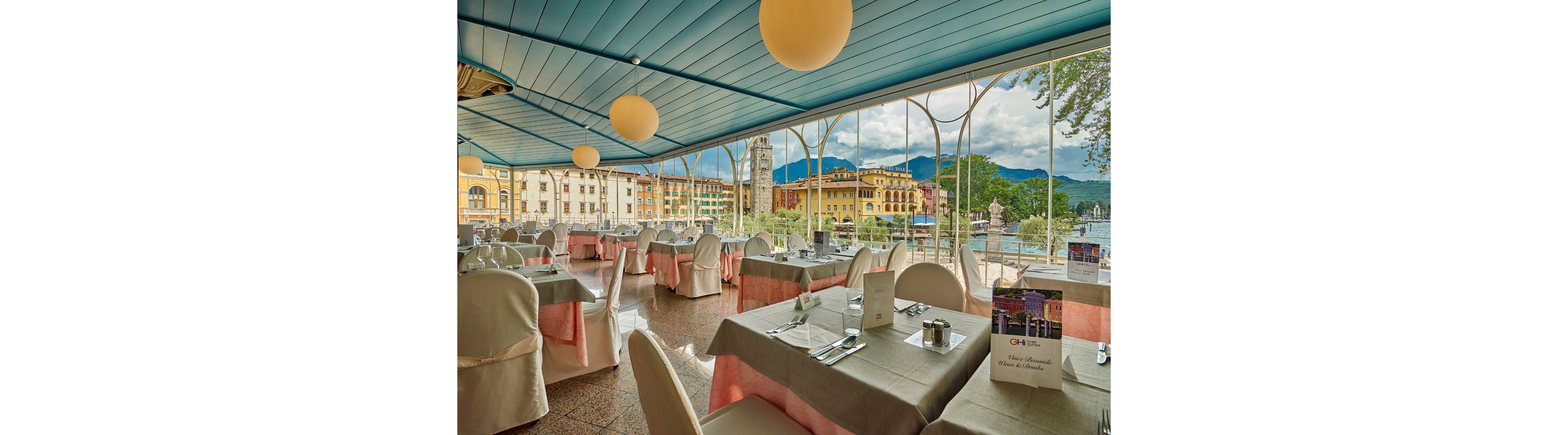 Spacious restaurant dining room with neatly arranged tables overlooks a picturesque lakeside town. Large windows frame colorful historic buildings, a tall clock tower, and distant mountains under a partly cloudy sky. Round pendant lights hang from a blue ceiling, and tables are covered with beige cloths and pink skirts, set with menus, cutlery, and condiments. The atmosphere feels calm and inviting, ideal for leisurely meals with a scenic waterfront view.
