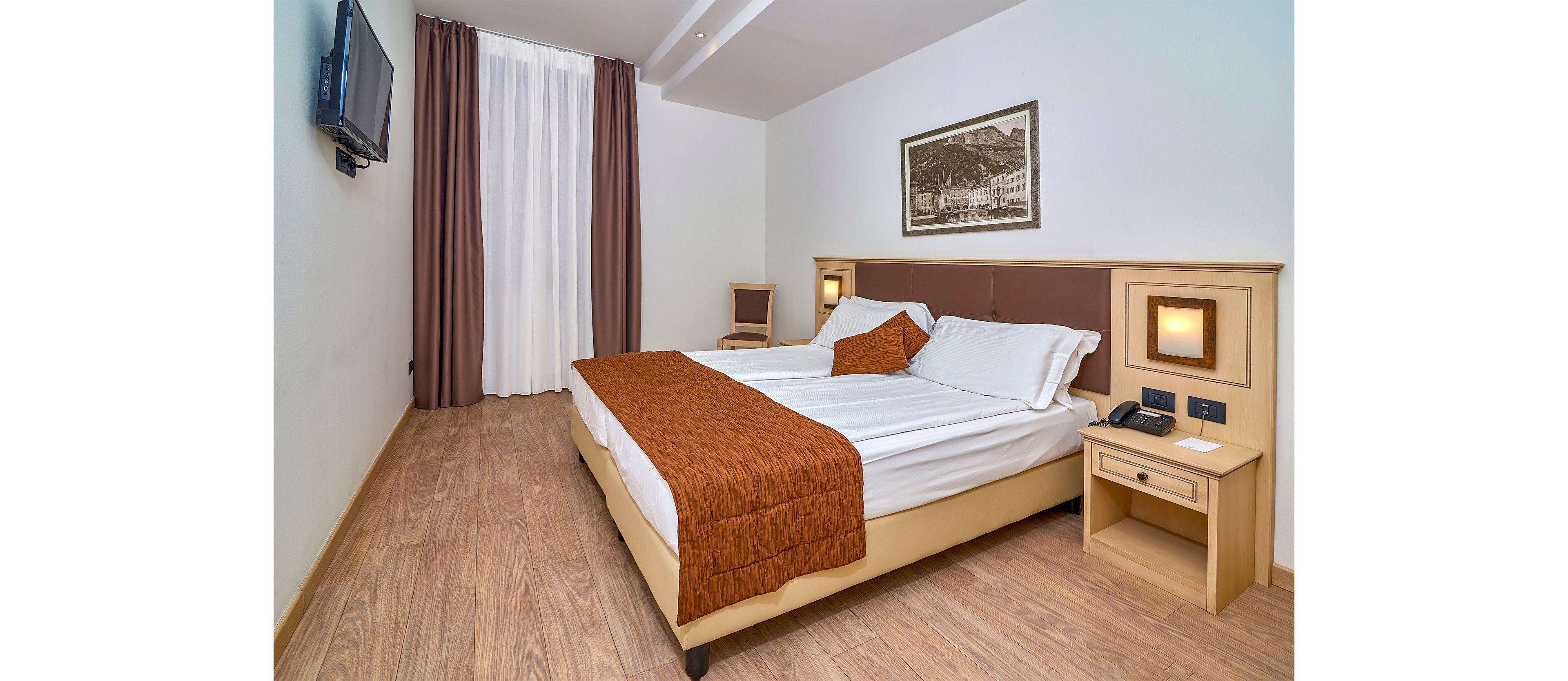 A clean, modern hotel room features a double bed with white linens and brown bed runners. The space includes wooden flooring, a bedside table with a telephone, and wall-mounted reading lights. A flat-screen television is mounted on the wall opposite the bed, and tall curtains cover a large window or glass door. A framed black-and-white picture hangs above the headboard, and a single chair sits in the corner, completing the simple, contemporary decor.