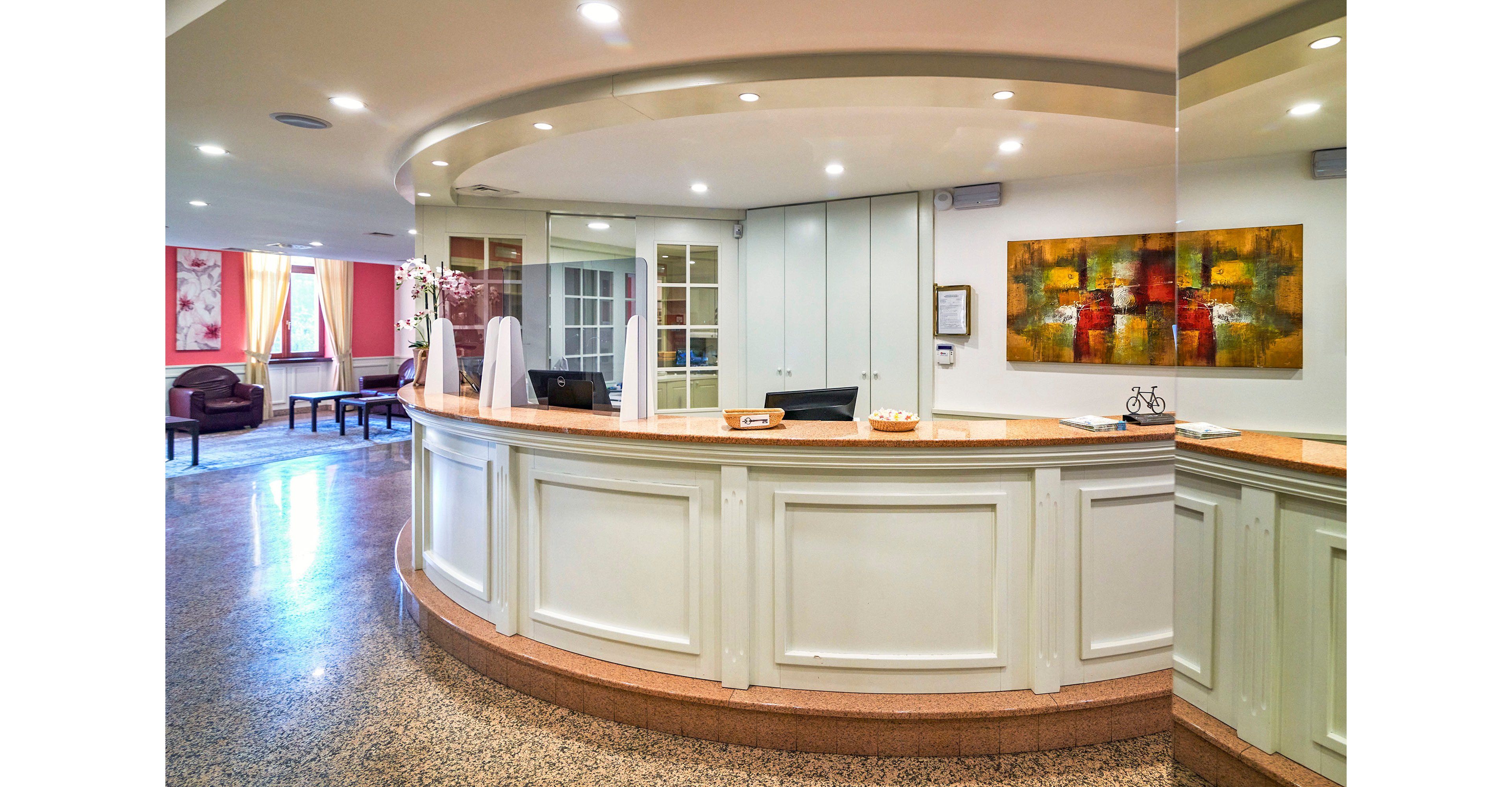 Curved white reception desk stands in a bright hotel lobby. The space features polished stone flooring, soft ceiling lighting, and large windows with pink curtains. A colorful abstract painting hangs on the wall behind the desk, and a vase of flowers adds a welcoming touch. Seating is visible in the background, creating an inviting atmosphere for guests.