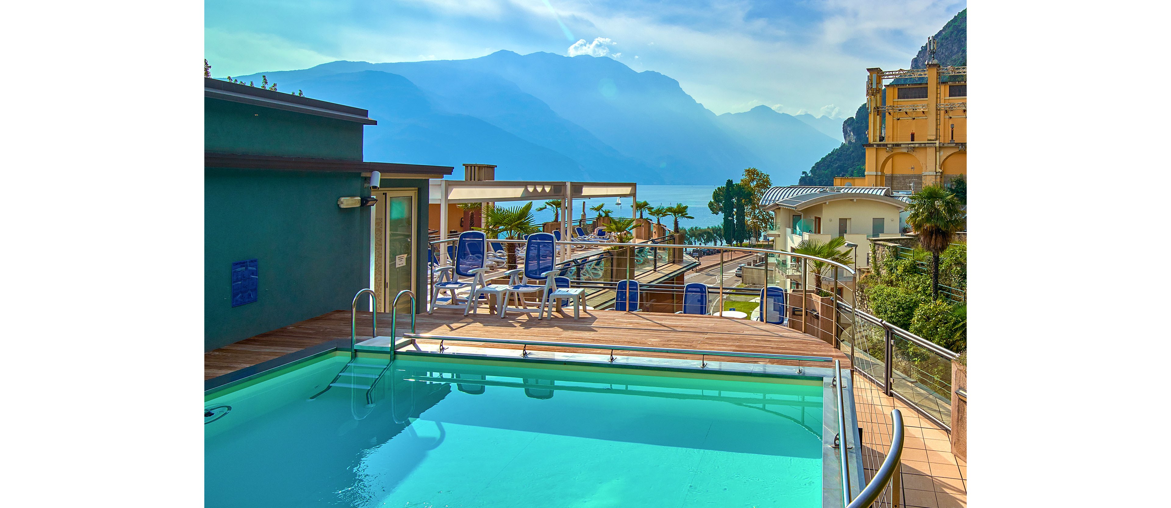 An outdoor rooftop swimming pool sits on a wooden deck with lounge chairs and umbrellas arranged around it. The terrace is part of a modern resort complex with multiple buildings and railings. In the background, a large lake is framed by hazy blue mountains under a bright, clear sky. Palm trees and greenery add a touch of tropical atmosphere to the scenic setting.