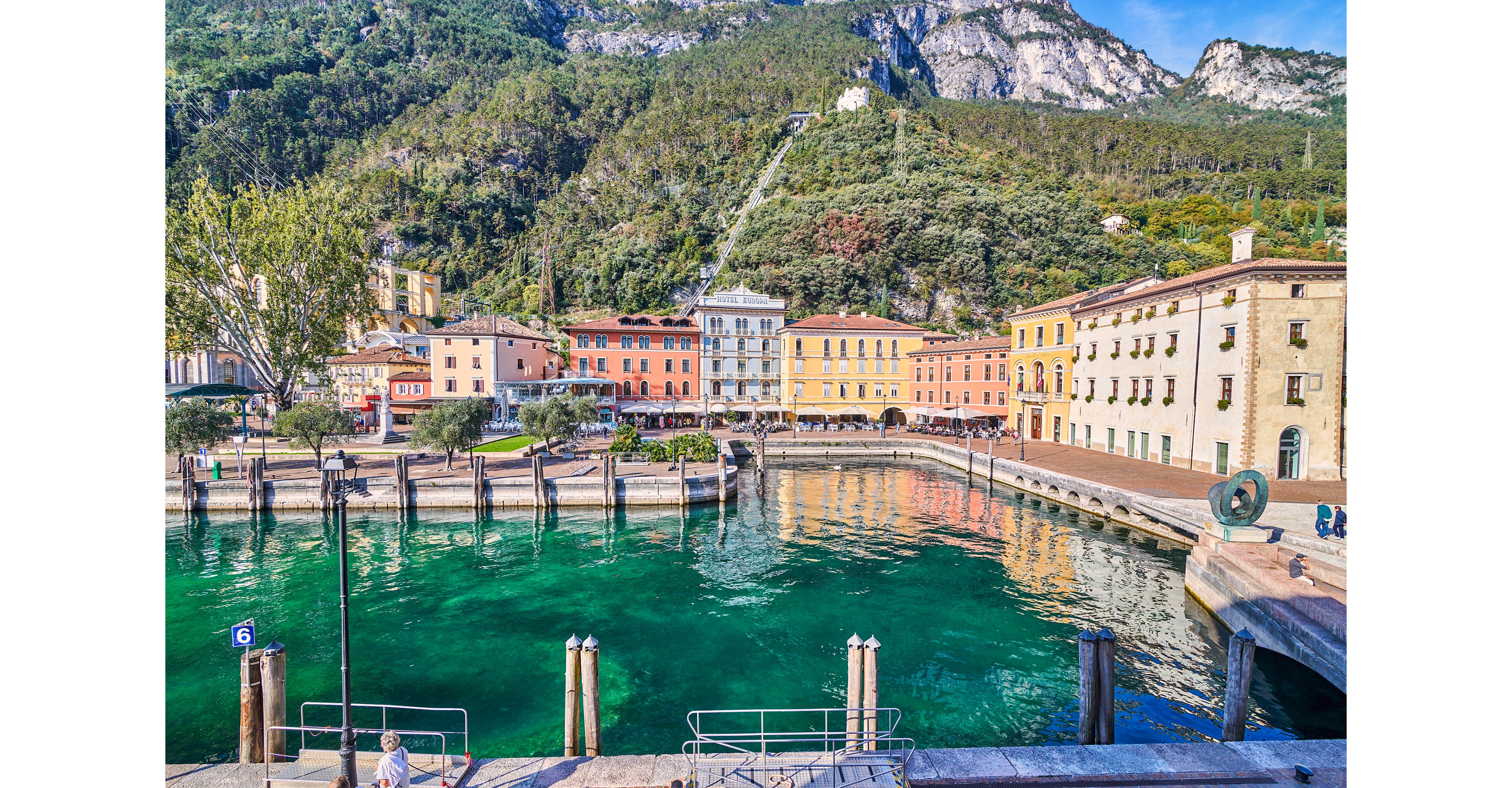 A vibrant lakeside town features colorful historic buildings lining the waterfront. Clear turquoise water reflects the facades and the surrounding mountainous landscape covered in greenery. Wooden piers and metal railings frame the harbor area, suggesting a quiet marina or docking zone. Bright daylight and crisp colors create a cheerful, inviting atmosphere in this scenic European-style setting.