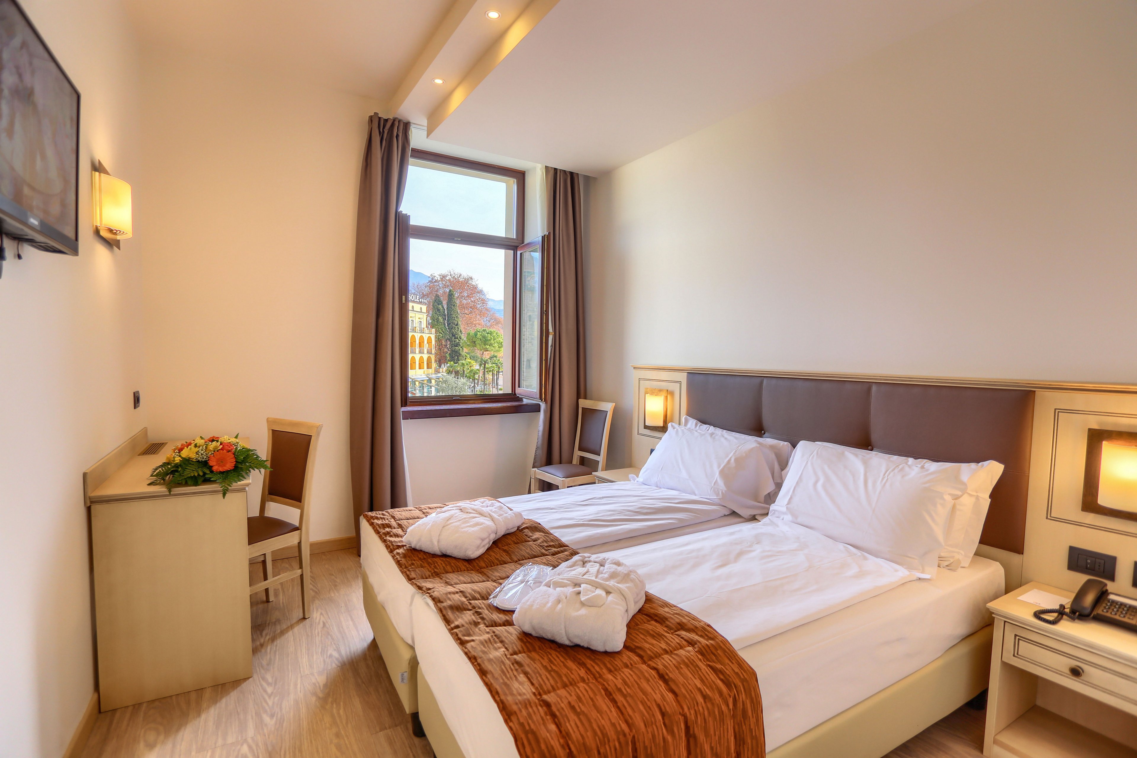 A neatly arranged hotel room features a large double bed with white linens and folded bathrobes at the foot. Warm lighting from wall sconces and a mounted television create a comfortable atmosphere. A desk with a chair and a bouquet of flowers sits near the window. Through the window, colorful buildings and trees are visible, adding a scenic touch to the interior space.