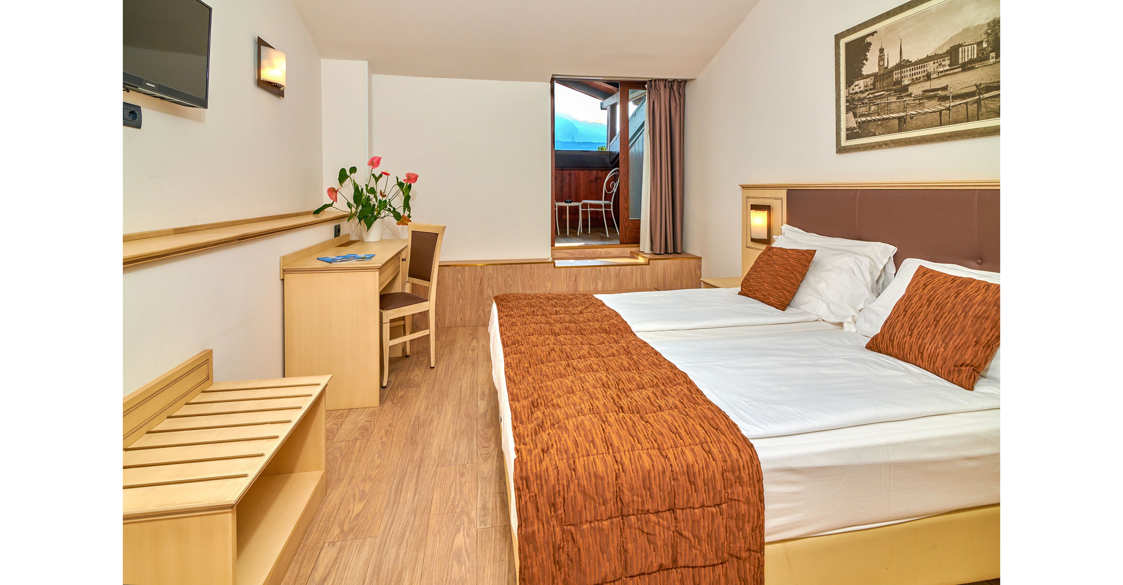 A tidy hotel room features two neatly made twin beds with white linens and rust-colored runners. The space includes light wood furniture, a small desk with a chair, and a vase of pink flowers adding a touch of color. A wall-mounted television and framed artwork decorate the room, while warm lighting creates a welcoming atmosphere. At the far end, an open doorway leads to a balcony with outdoor chairs and a scenic mountain view.