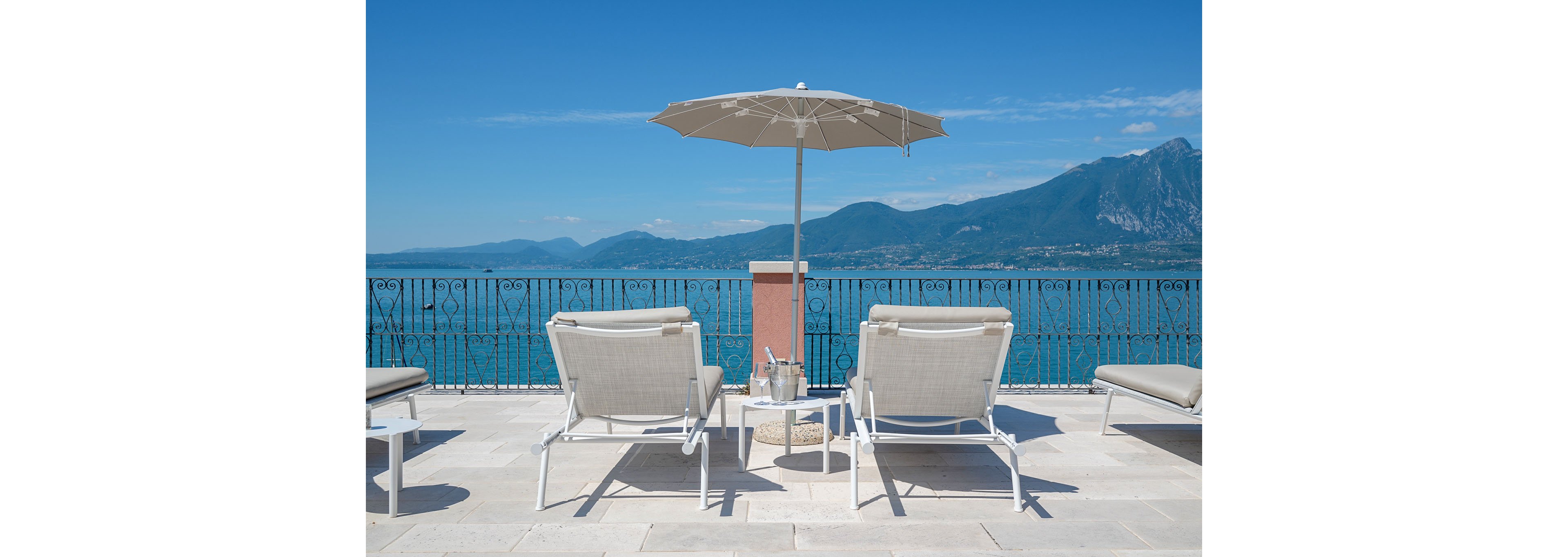 Two white sun loungers sit on a bright stone terrace facing a calm blue lake and distant mountains. A tall beige umbrella provides shade between the chairs, with a small side table holding glass bottles and tumblers. The wrought-iron railing frames the scenic waterfront view under a clear, cloudless sky. The scene suggests a peaceful vacation setting ideal for relaxation and sunbathing.
