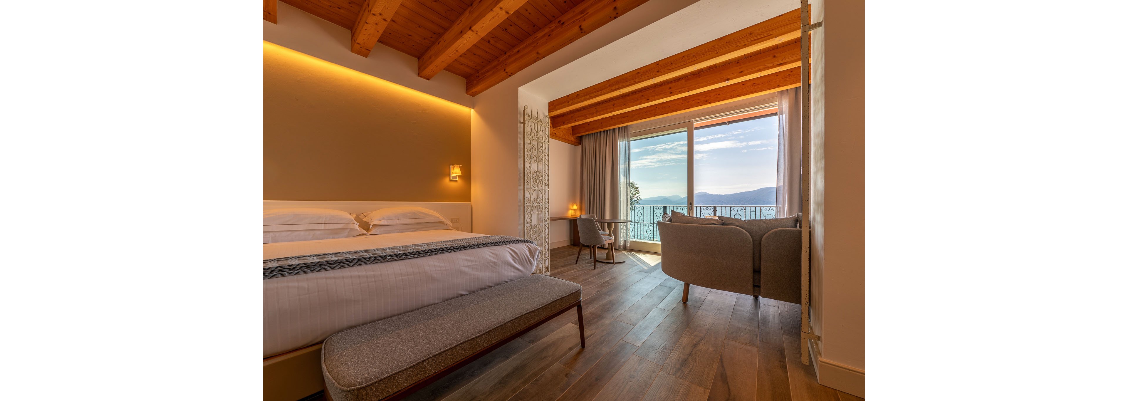 A modern hotel bedroom features a large neatly made bed with a cushioned bench at the foot. Warm wooden beams and flooring create a cozy atmosphere, complemented by soft wall lighting. At the far end of the room, a seating area with two armchairs faces a large window and glass door. Beyond the balcony railing, a scenic view of mountains and water is visible under a bright sky.