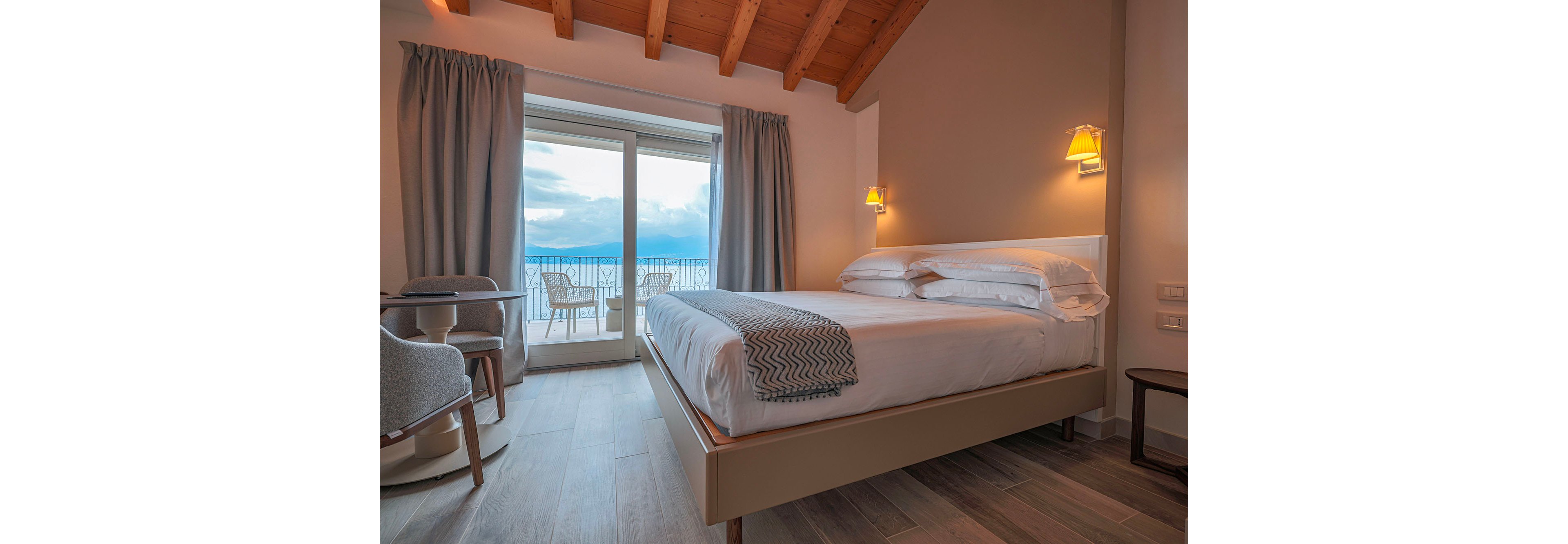 A neatly made double bed with white linens sits in a modern hotel bedroom. Warm wall sconces and a wooden beamed ceiling create a cozy atmosphere. Large glass doors open onto a balcony with chairs, overlooking a cloudy sky and distant water. Neutral tones, soft curtains, and contemporary furniture contribute to a calm, inviting interior.