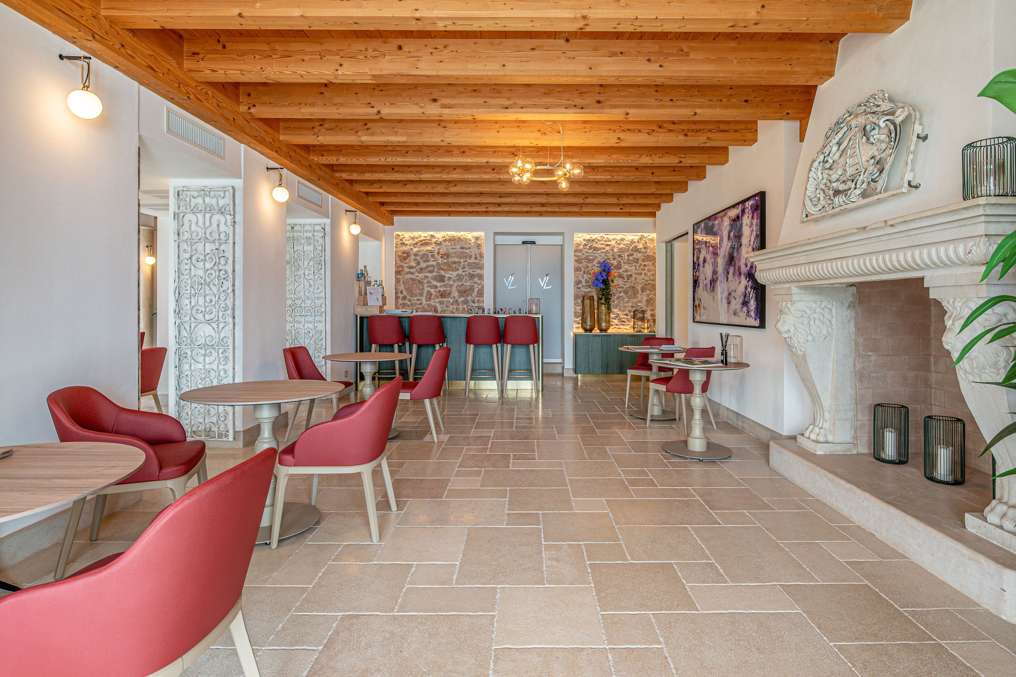 A contemporary lounge area features round wooden tables paired with red upholstered chairs. The space has a light stone tile floor and a rustic wooden beam ceiling, creating a warm and inviting atmosphere. Decorative elements include a large white fireplace, patterned room dividers, and colorful wall art. Soft ambient lighting and a small bar counter at the back enhance the relaxed, modern interior design.