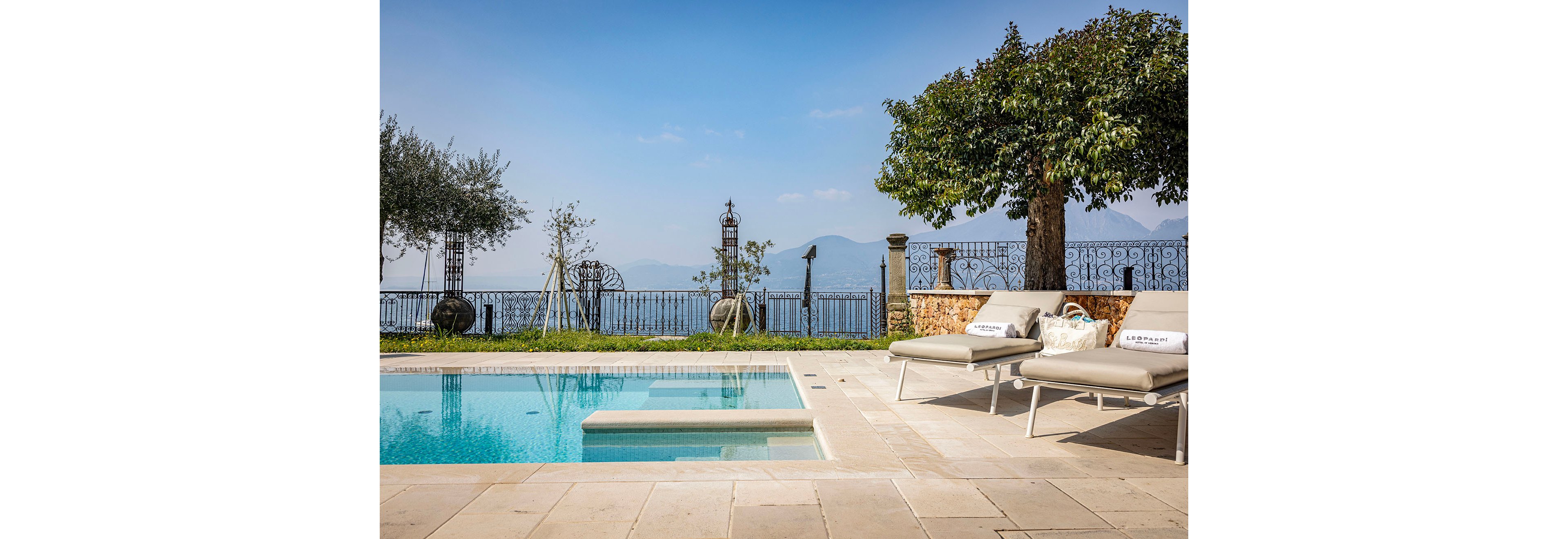 A serene outdoor swimming pool sits on a stone terrace with two cushioned lounge chairs arranged beside it. Beyond the pool, an ornate metal fence frames a panoramic view of a distant lake and hazy mountains under a clear blue sky. Mature trees and potted plants add greenery and shade to the elegant setting. The scene evokes a calm, luxurious vacation atmosphere in a private villa or upscale resort.