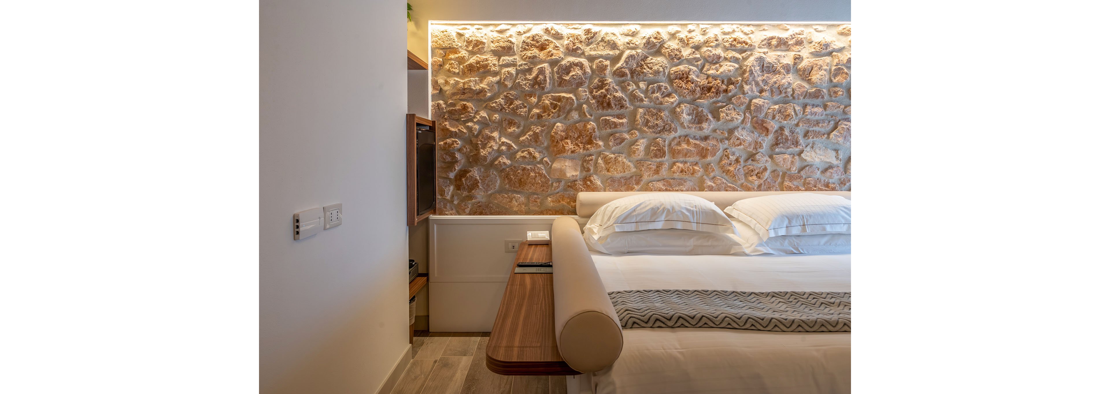 A modern bedroom features a neatly made bed with stacked white pillows and a light-colored headboard. Behind the bed, a textured stone accent wall is highlighted by warm recessed lighting, creating a cozy atmosphere. A wooden side table extends from the bed frame, holding a small device and adding functional style. The room combines natural materials and soft lighting for a calm, contemporary interior design.