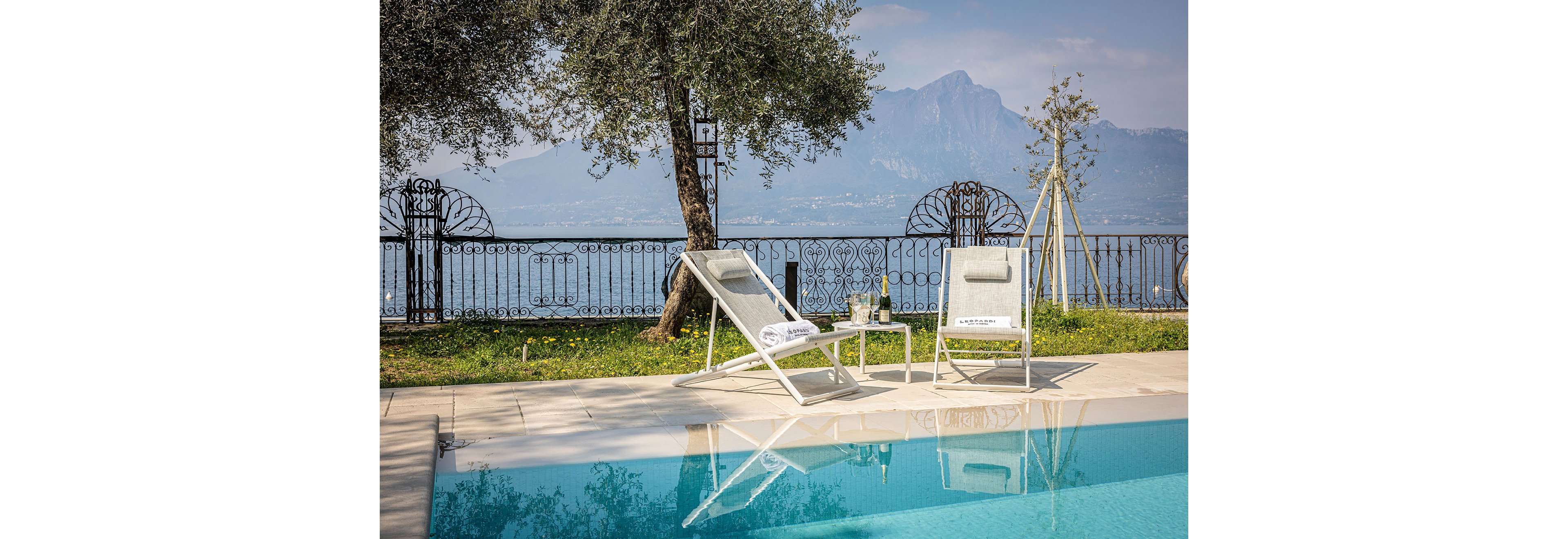 Two white lounge chairs sit beside a clear blue swimming pool, facing a calm lake and distant mountains. A small side table holds a bottle and glasses, suggesting a relaxing outdoor refreshment. Ornate metal railings and trees frame the view, adding elegance to the lakeside terrace. The scene feels tranquil and leisurely, with soft natural light and reflections in the pool water.