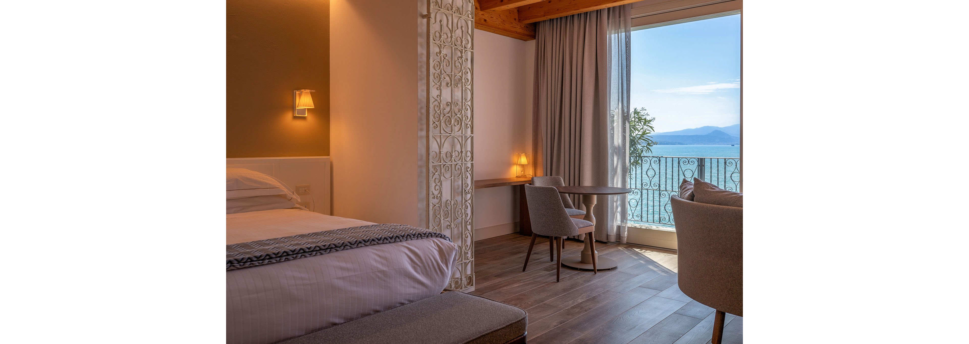 A neatly made bed sits in a cozy hotel room with warm lighting and wooden ceiling beams. A small table and two chairs are positioned near a large window that opens onto a balcony. Sheer curtains frame the view of the sea and distant mountains outside. Soft neutral tones and natural light create a calm, inviting atmosphere ideal for relaxation.
