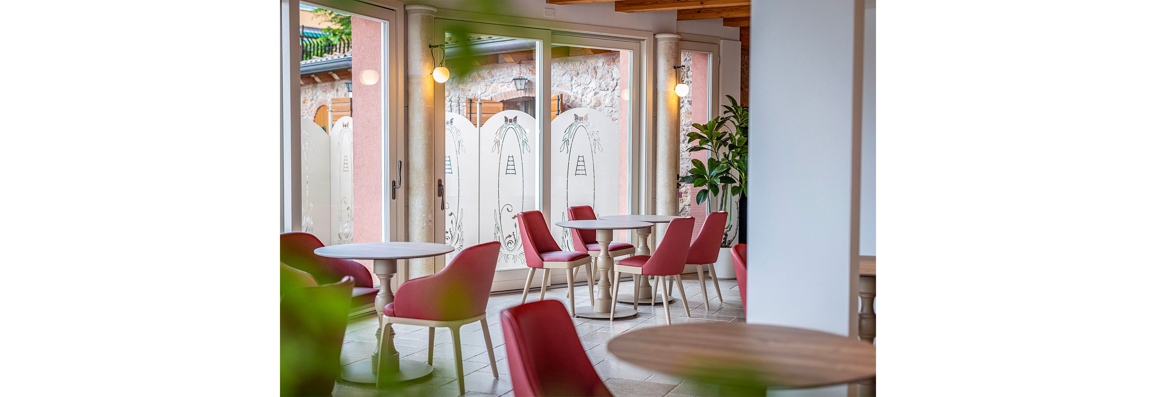 A modern cafe interior features round wooden tables paired with red upholstered chairs. Large glass doors open to an outdoor area, allowing natural light to fill the space. Decorative panels with simple line drawings and wall-mounted lights add visual interest. Green plants and warm wooden ceiling elements contribute to a cozy, inviting atmosphere.