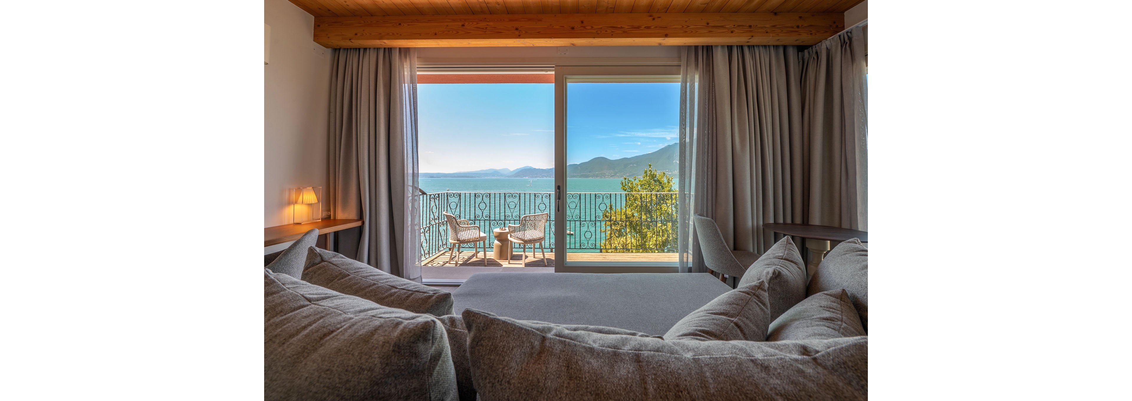 A cozy hotel room features a large bed or sofa facing a sliding glass door that opens onto a balcony. Two wicker chairs and a small round table sit on the balcony, overlooking a calm blue sea and distant mountains. Soft gray curtains frame the doorway, and warm wooden ceiling details add to the inviting atmosphere. The scene suggests a peaceful vacation setting with bright natural daylight and a relaxing coastal view.