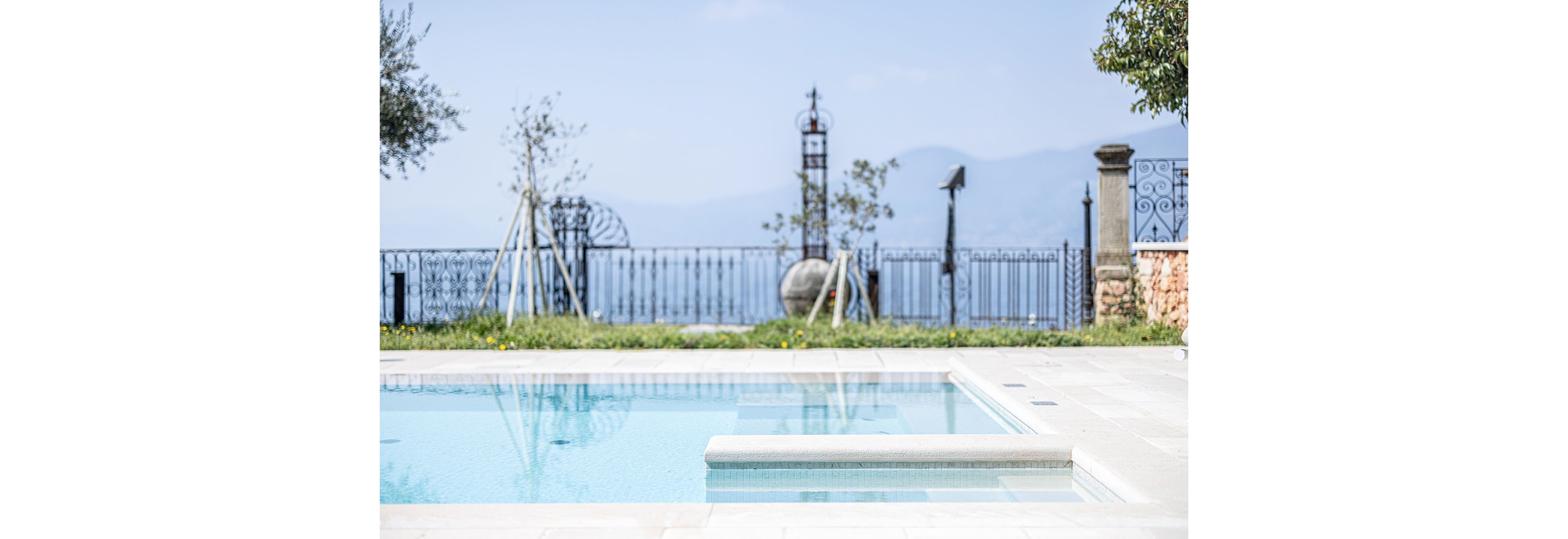 A clear blue swimming pool sits in the foreground of an outdoor terrace. Beyond the pool, a decorative metal fence lines the edge of the property, overlooking distant mountains and a hazy sky. Young trees and sculptural garden elements add interest along the fence line. The bright daylight and soft colors create a calm, luxurious vacation atmosphere.