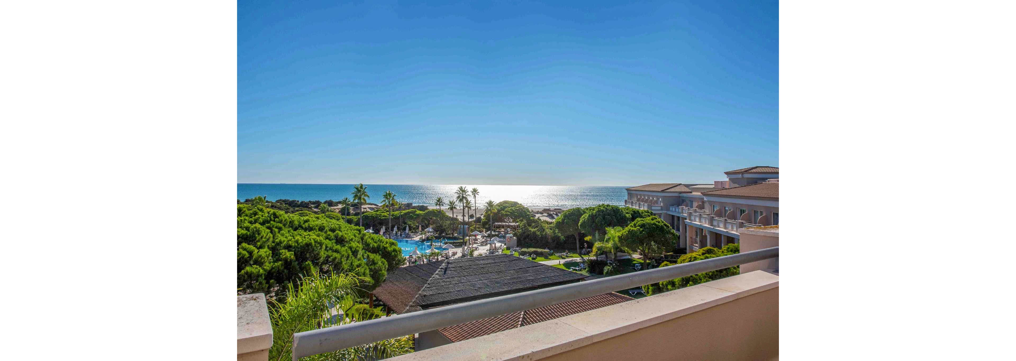 A bright seaside resort is seen from a balcony overlooking lush green trees and hotel buildings. The sun reflects off the calm ocean, creating a shimmering horizon under a clear blue sky. Below, a swimming pool area and palm trees suggest a relaxing vacation atmosphere. The composition emphasizes the open balcony railing in the foreground leading the eye toward the distant water.