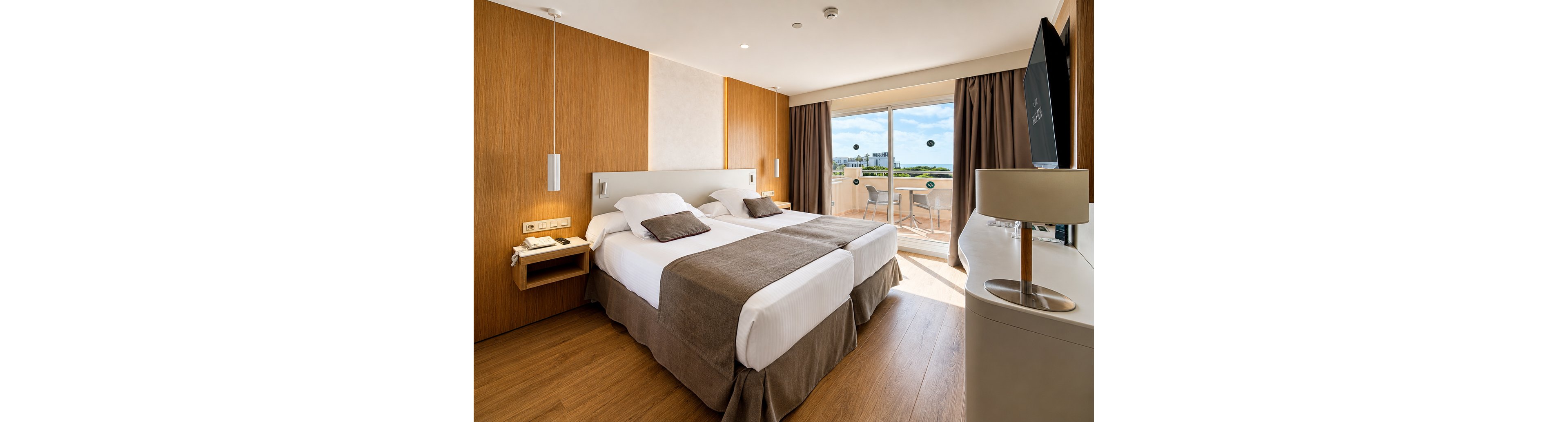 A contemporary hotel room features two neatly made twin beds with white linens and brown throws. Warm wooden walls and flooring create a cozy, minimalist atmosphere, complemented by simple bedside lighting and a wall-mounted television. Large sliding glass doors open to a balcony with outdoor seating and a scenic view of greenery under a blue sky with clouds. The space appears bright, clean, and inviting, ideal for comfortable short stays or vacations.