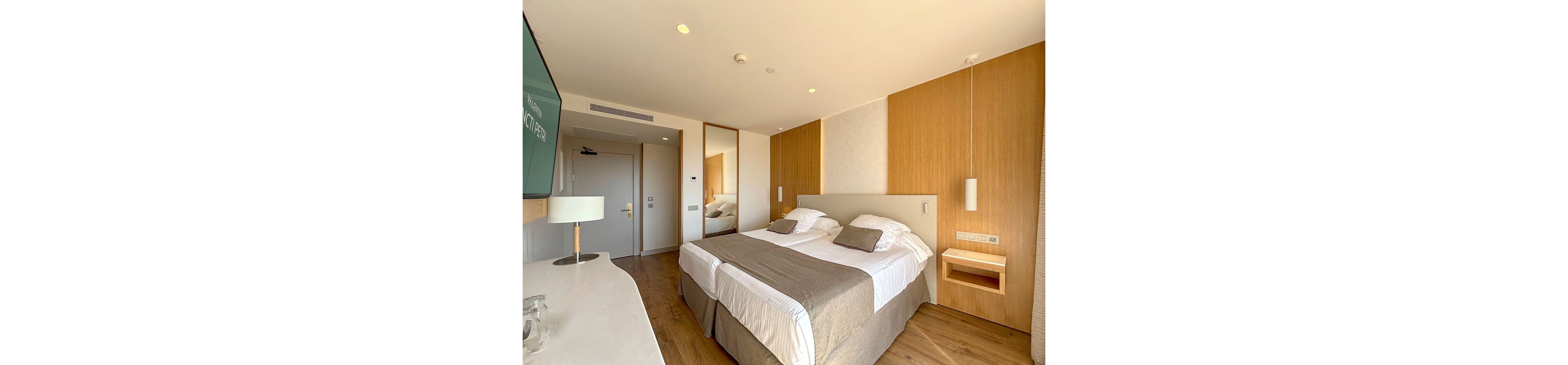 A contemporary hotel room features a large double bed with white linens and beige throws. The space is decorated in warm wood tones, with built-in bedside shelves and minimalist lighting. A flat-screen television is mounted on the wall opposite the bed, and a desk area runs along the side. Natural light floods the room through an unseen window, creating a clean and inviting atmosphere.