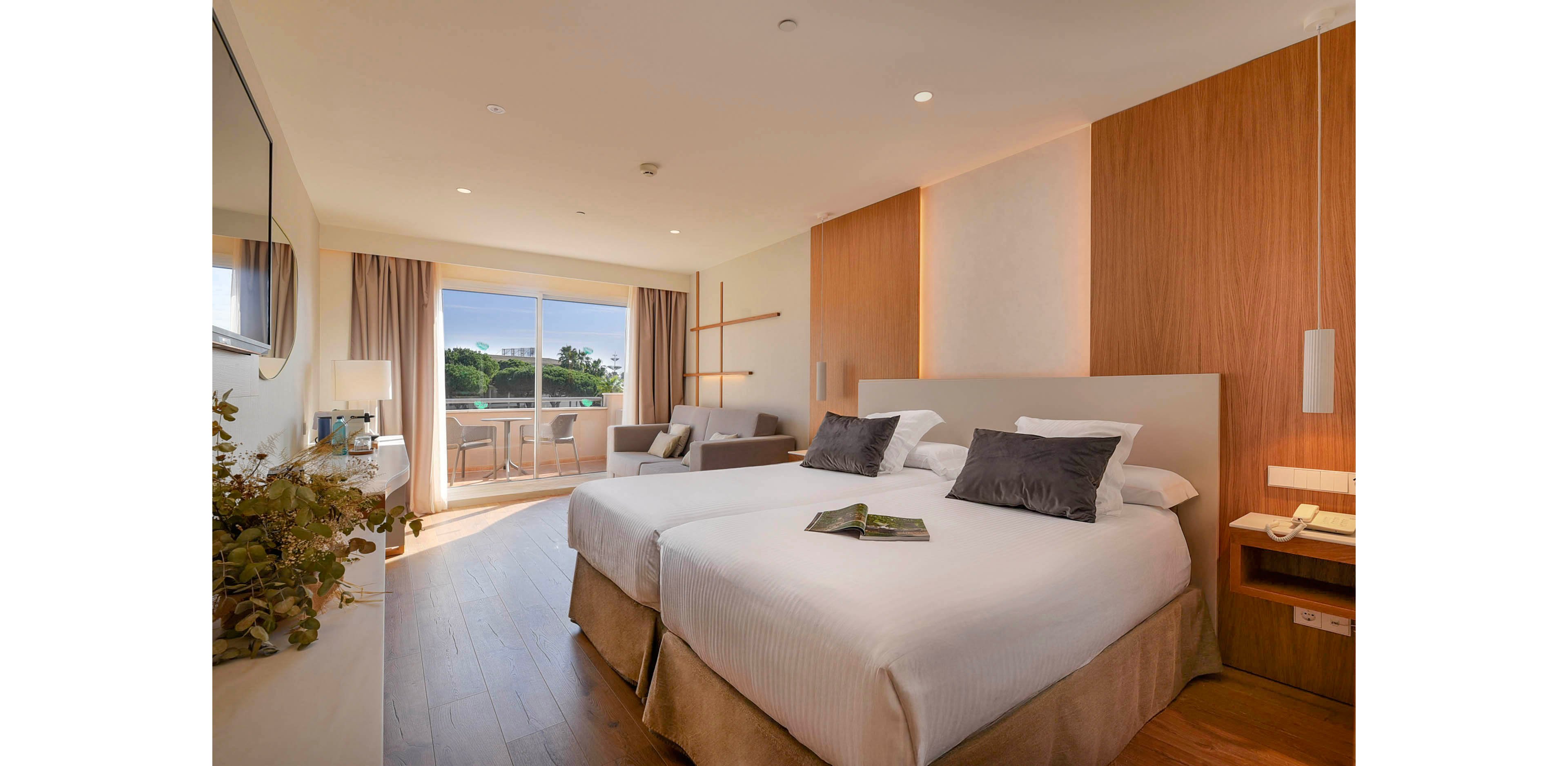 A contemporary hotel room features two neatly made twin beds with white linens and dark accent pillows. The space includes a small sitting area with a sofa and coffee table near a large sliding glass door. Warm wooden paneling and flooring create a cozy, minimalist atmosphere. Natural light fills the room, and a balcony with outdoor seating overlooks greenery outside.
