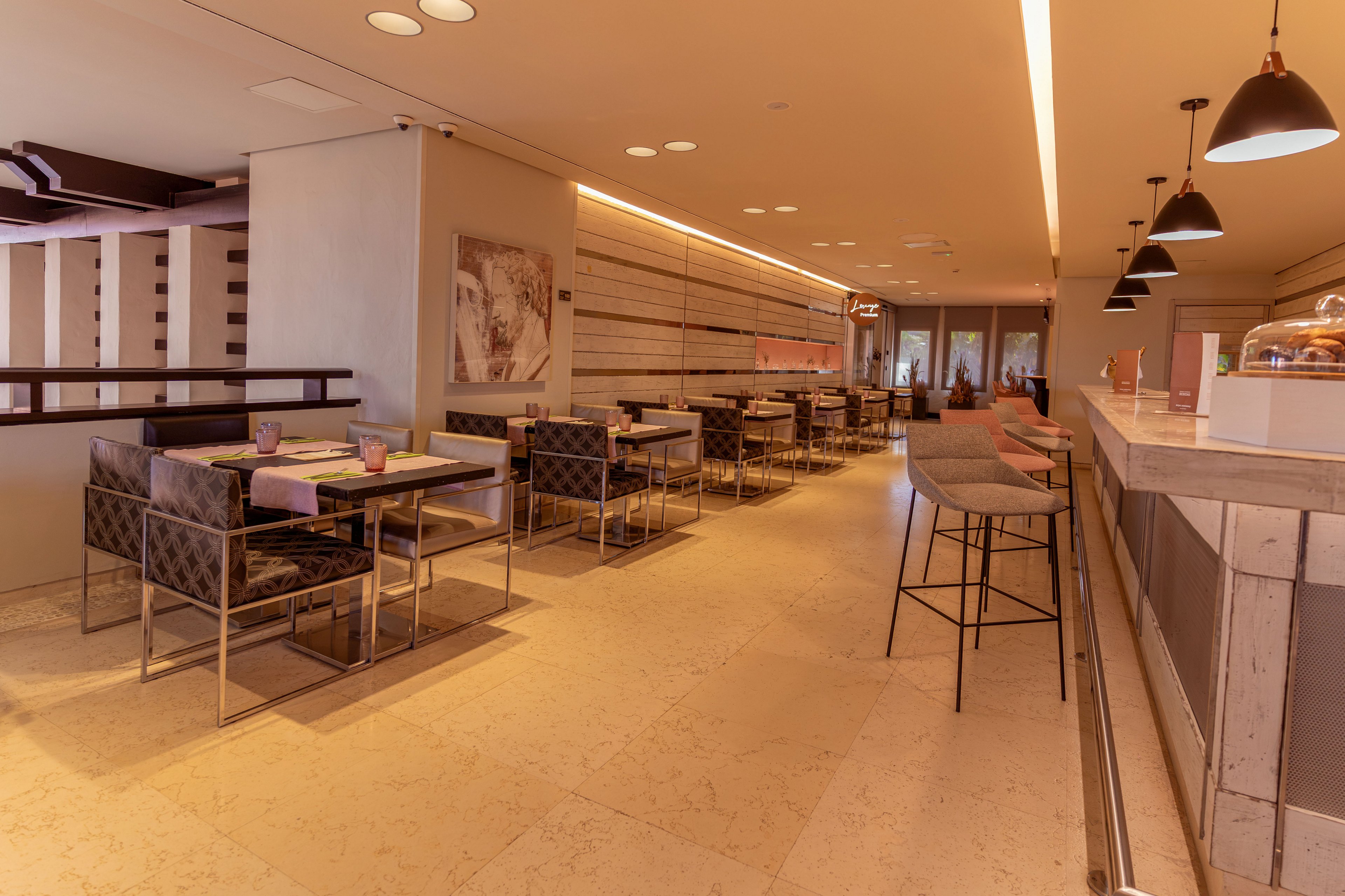 A contemporary cafe interior features neatly arranged tables and cushioned chairs along one side of the room. A long counter with high stools runs parallel to the seating area, creating a casual bar-style space. Warm ambient lighting and neutral tones give the cafe a cozy, inviting atmosphere. The room appears clean and empty, with no people present and large windows at the far end providing natural light.