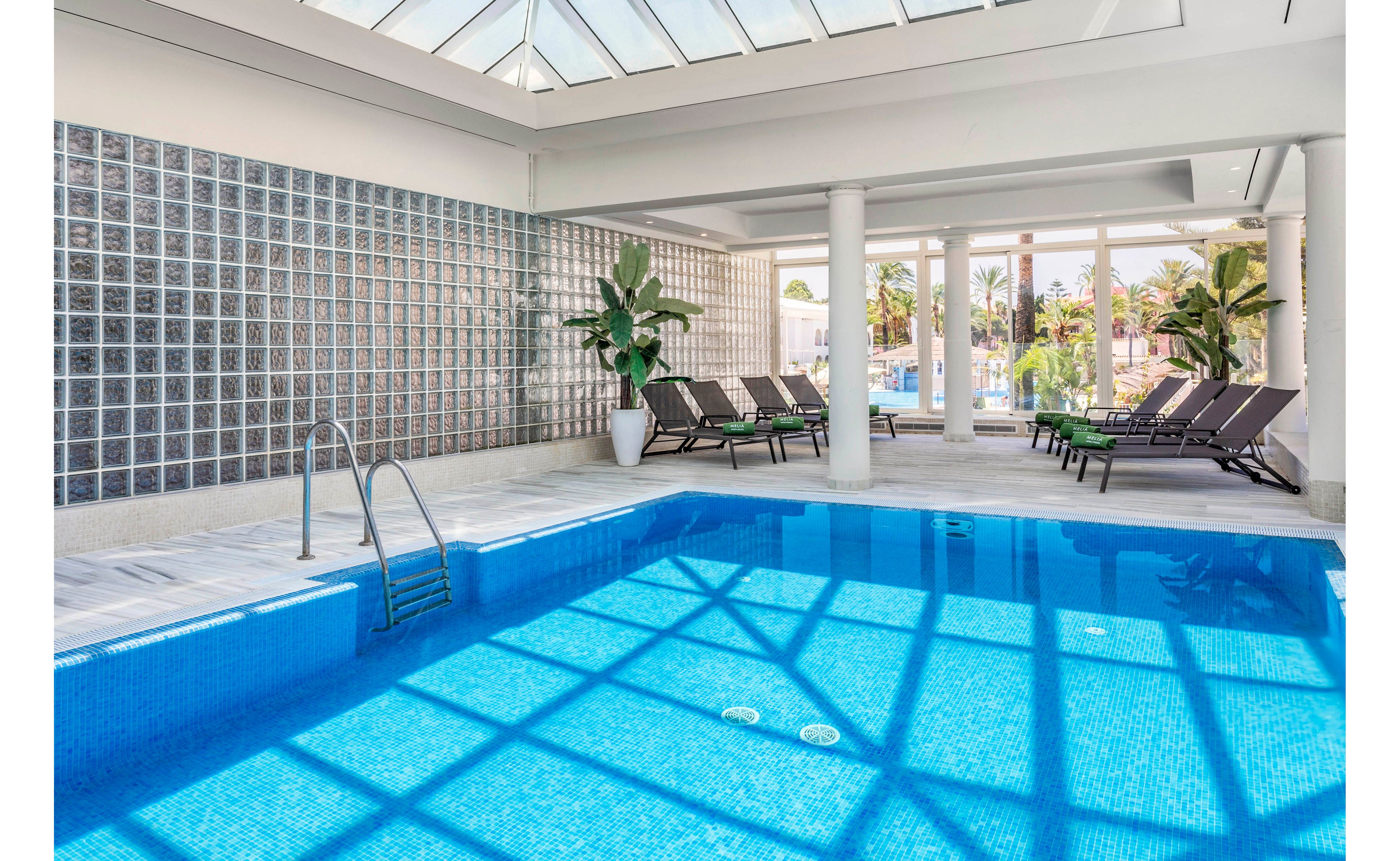 A clean indoor swimming pool is shown in a bright, sunlit space. Glass block walls and large skylights allow natural light to cast geometric shadows across the blue water. Several dark lounge chairs line the poolside near tall potted plants, creating a relaxing resort atmosphere. Through the windows, an outdoor area with palm trees and additional seating is visible.