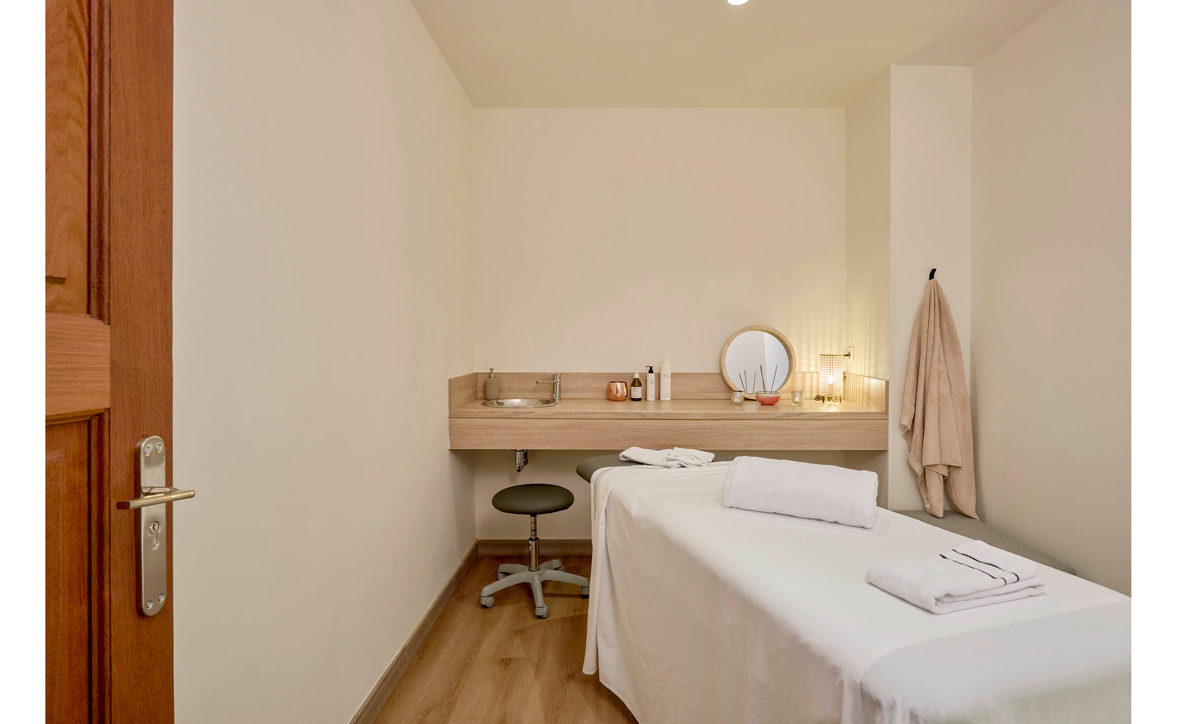 A clean, minimalist spa treatment room features a neatly made massage table with folded white towels. A wooden counter at the back holds skincare bottles, a round mirror, and decorative items, creating a calm atmosphere. A beige robe hangs on the wall, and a small stool sits under the counter, suggesting a professional therapist workspace. Neutral colors and soft lighting enhance the relaxing, wellness-focused setting.