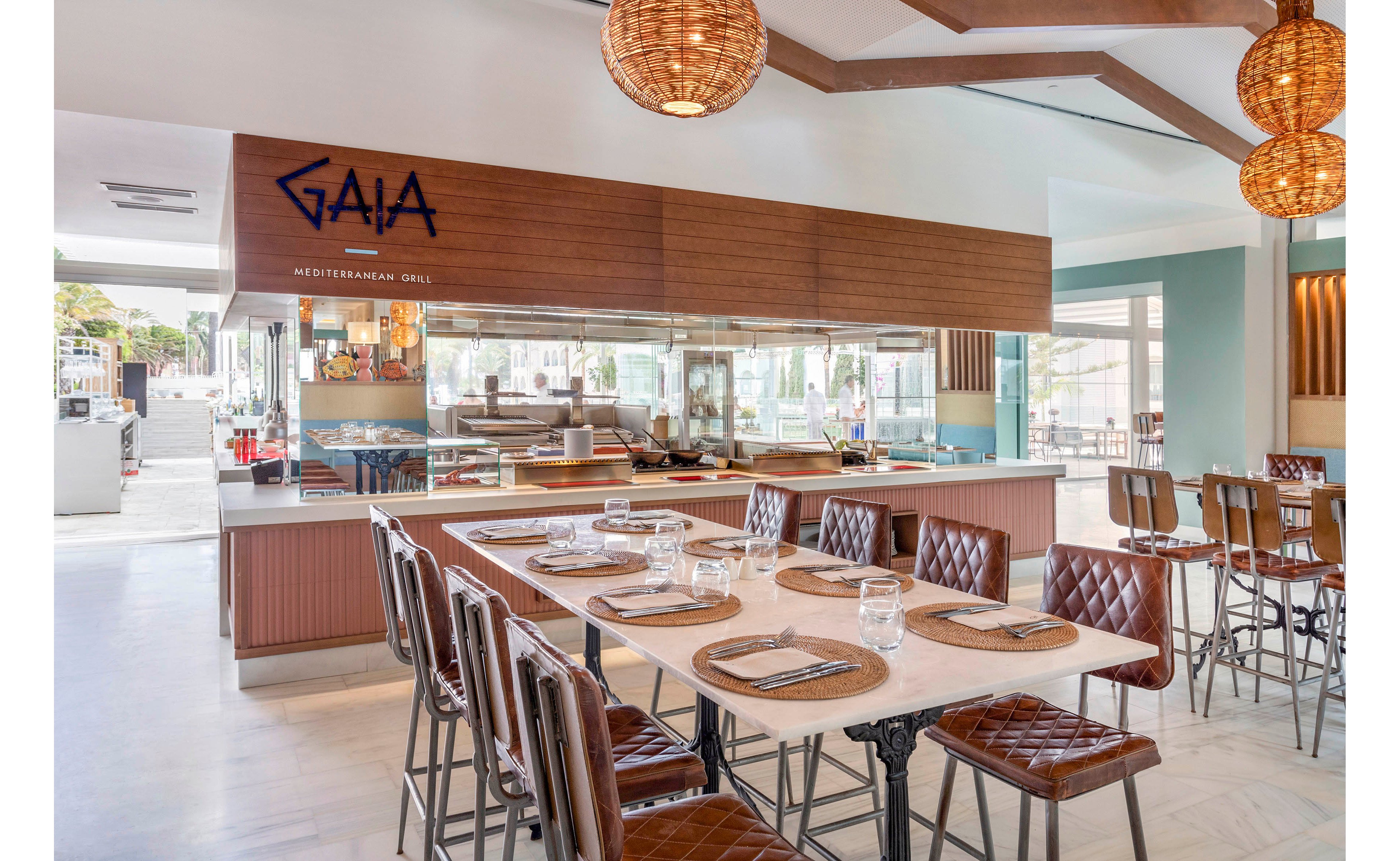 The image shows a contemporary restaurant interior featuring the unmistakable 'GAIA Mediterranean Grill' sign above the serving counter. The setting includes neatly arranged tables with brown leather chairs, glassware, and woven pendant lights, creating a warm and inviting atmosphere. Staff members are visible behind the counter, preparing food in an open kitchen. The overall palette is neutral with wood accents and soft lighting, emphasizing a relaxed dining experience.
