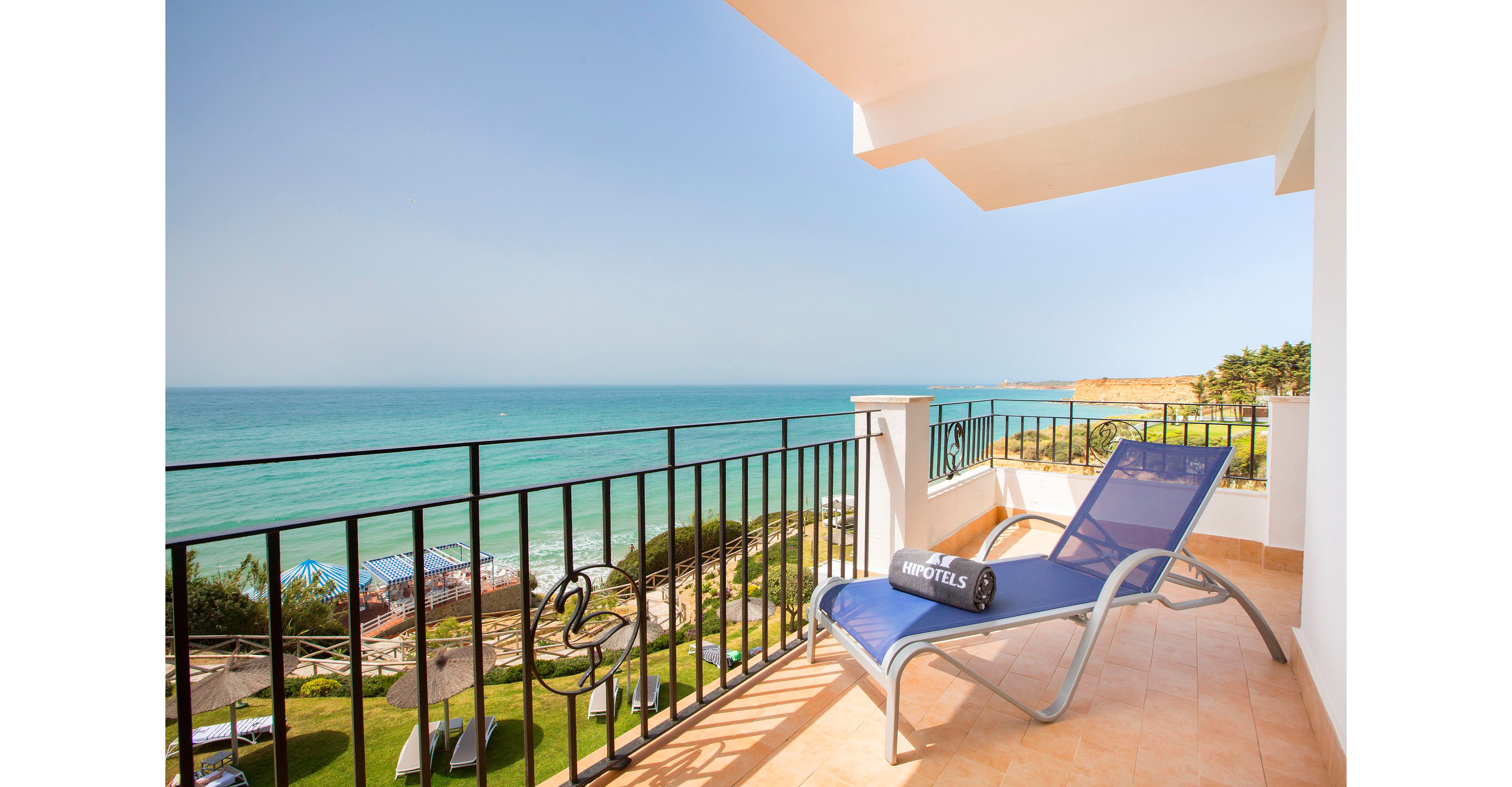 A bright hotel balcony features a blue lounge chair and towel facing a wide expanse of turquoise ocean. The scene looks out over a coastal area with beach umbrellas, greenery, and a distant cliff under a clear blue sky. Warm tiled flooring and black railings frame the relaxing seaside view. The overall atmosphere is calm, sunny, and inviting for a beach vacation.