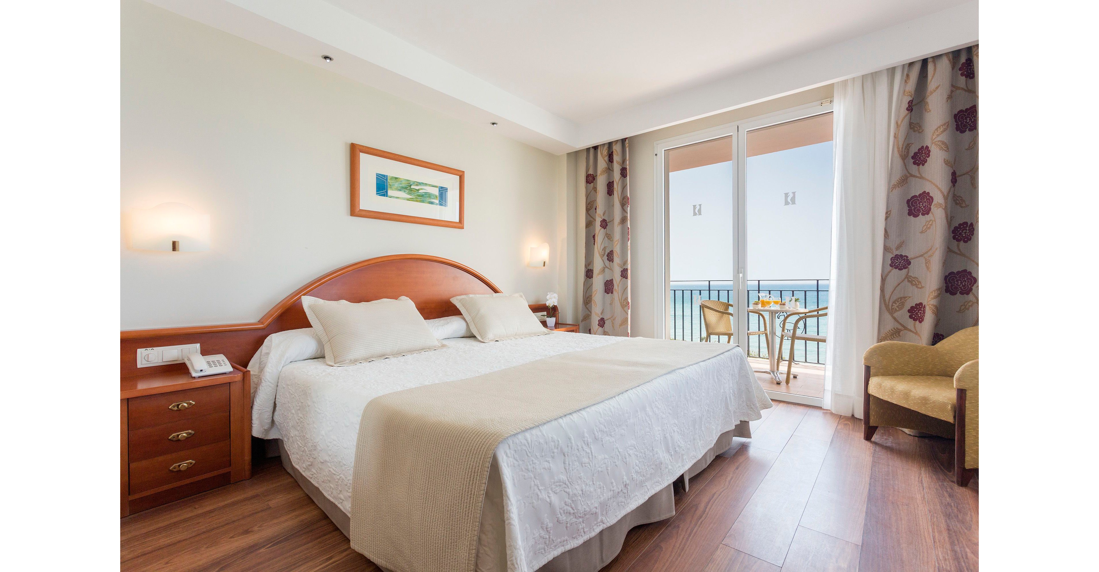 A neatly made double bed with neutral bedding is the focal point of a bright hotel room. Wooden furniture, including bedside tables and an armchair, adds warmth to the contemporary interior. Large glass doors open onto a balcony with a table and chairs, overlooking the sea. Soft lighting and simple wall art create a calm, inviting atmosphere ideal for relaxation.