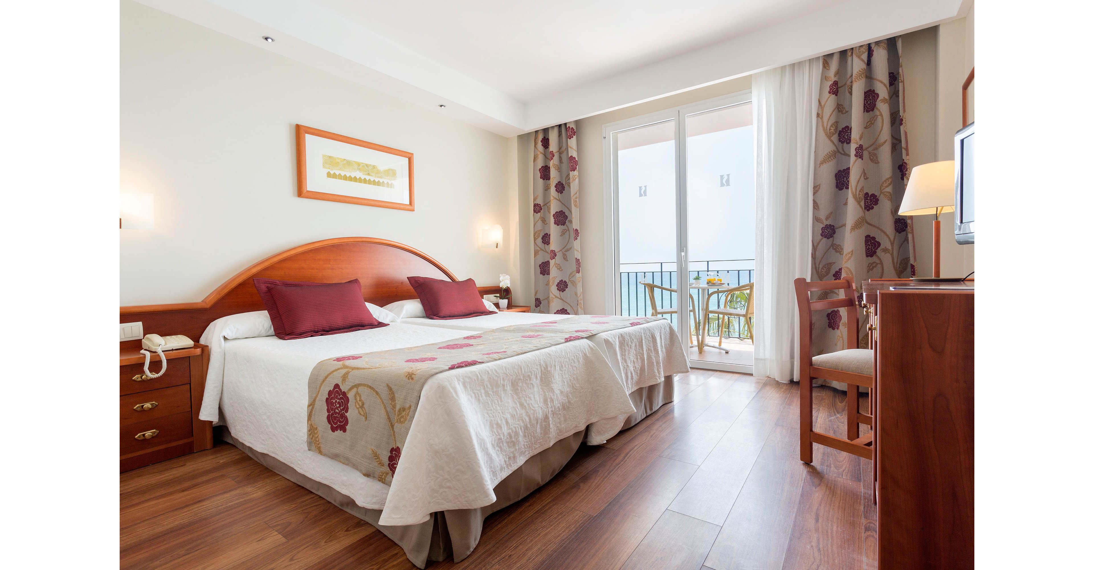 A tidy hotel bedroom features a double bed with white linens, floral bed runner, and red accent pillows. Warm wooden furniture, including bedside tables, a desk, and a chair, complement the light walls and hardwood floor. Large glass doors open to a small balcony with outdoor chairs and a table, letting in natural daylight. Patterned curtains and a framed artwork above the bed add subtle decorative touches to the cozy space.