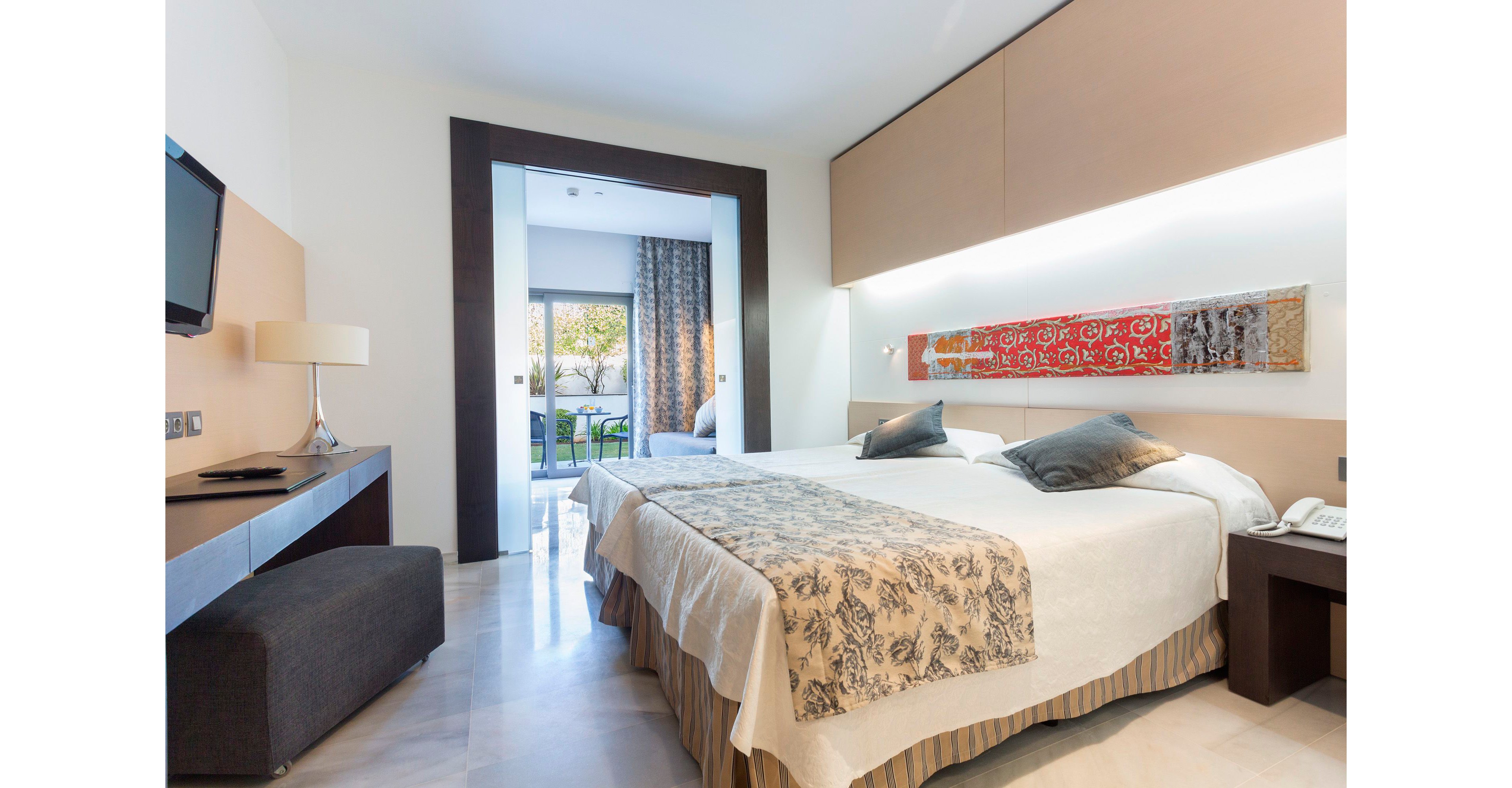 A contemporary hotel room features a large double bed with patterned runner and accent pillows. The space is bright and minimalist, with light-colored walls, sleek furniture, and a wall-mounted television. A decorative horizontal artwork adds color above the bed, while a sliding doorway opens to a terrace with outdoor seating and greenery. Natural light floods the room, creating an inviting and comfortable atmosphere for guests.