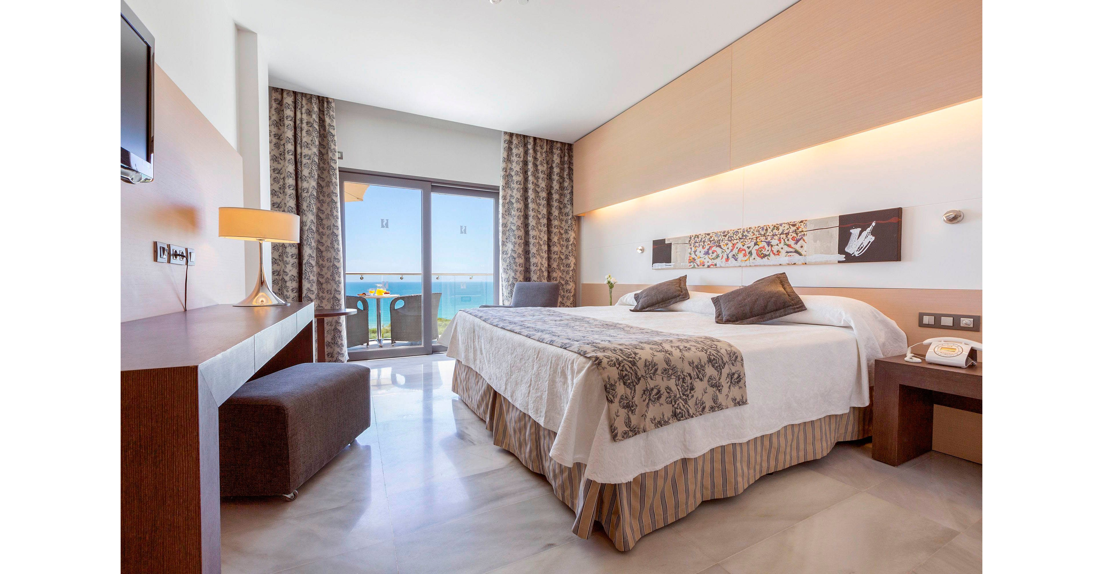 A bright, modern hotel bedroom features a large double bed with patterned runner and accent pillows. Floor-to-ceiling curtains frame a glass door that opens to a balcony overlooking the ocean. A sleek wooden desk, upholstered bench, and bedside tables with lamps complete the contemporary furnishings. Neutral tones and soft lighting create a calm, inviting atmosphere ideal for relaxation.