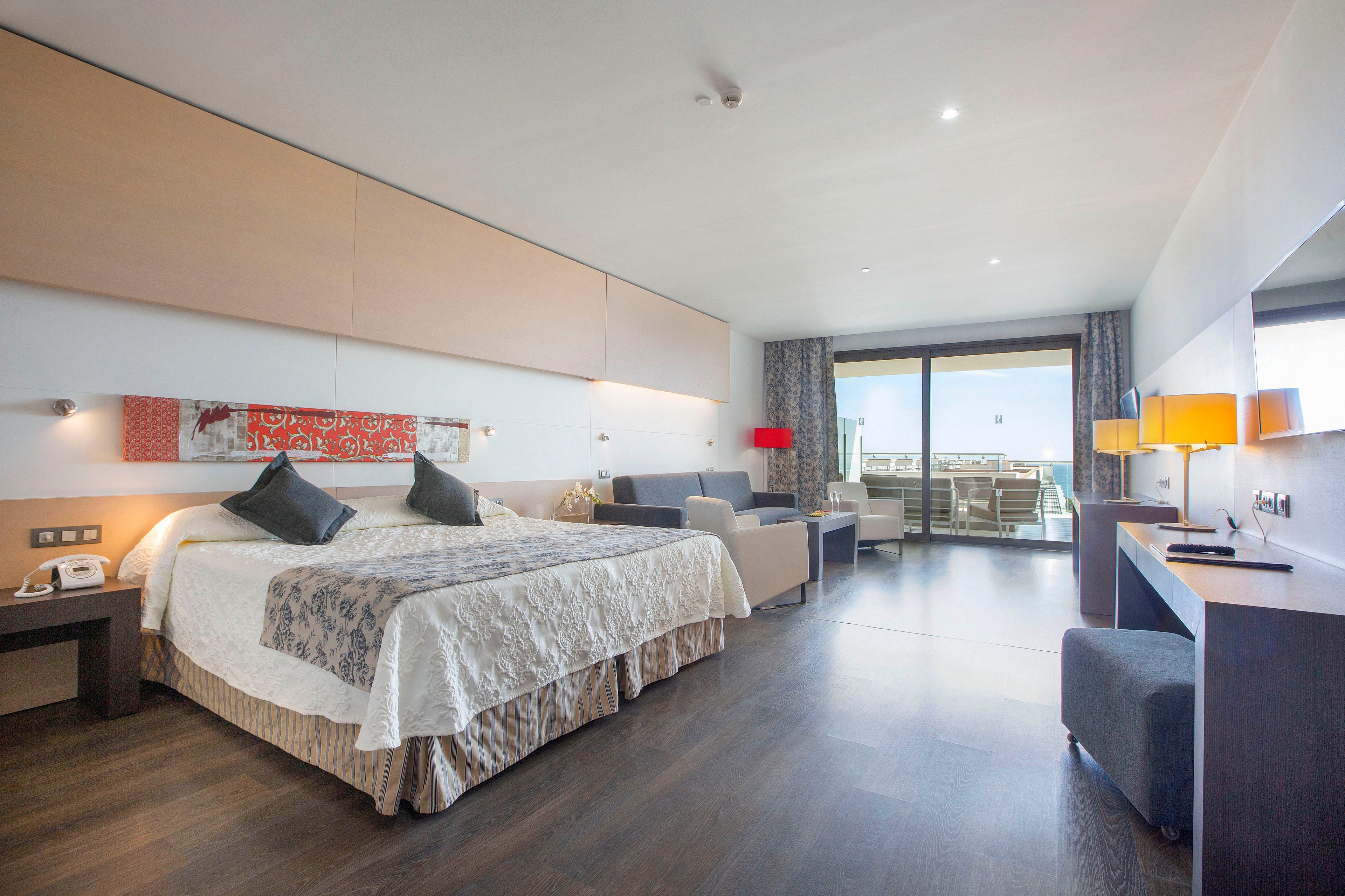 Spacious contemporary hotel room featuring a large bed with decorative pillows and a patterned throw. The room includes a sitting area with a sofa, side tables, and accent lamps in warm colors. A work desk and chair are positioned along the wall, complementing the sleek wooden flooring. Floor-to-ceiling glass doors open onto a balcony with outdoor seating and a distant view.