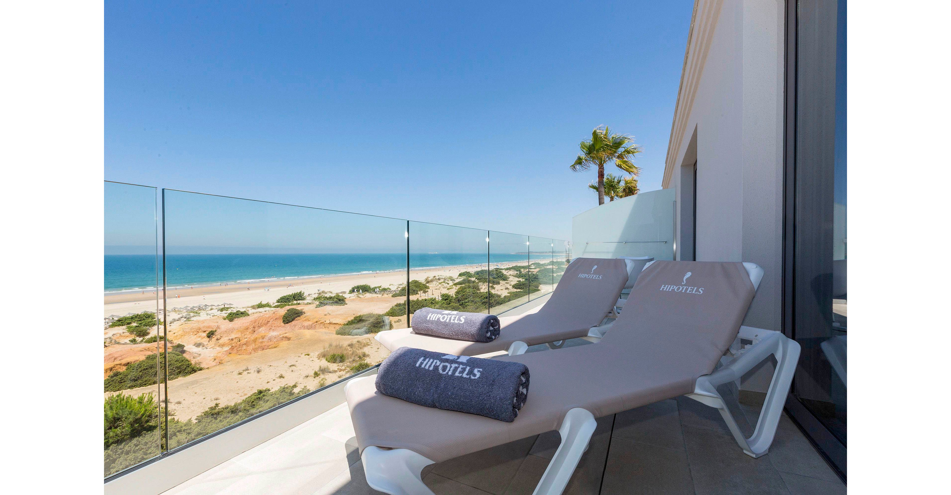 Two beige sun loungers with rolled towels are arranged on a modern balcony overlooking a sandy coastline. A clear glass railing provides an unobstructed view of the beach, dunes, and blue ocean. The sky is bright and cloudless, creating a calm and relaxing atmosphere. A single palm tree adds a tropical touch to the contemporary hotel setting.