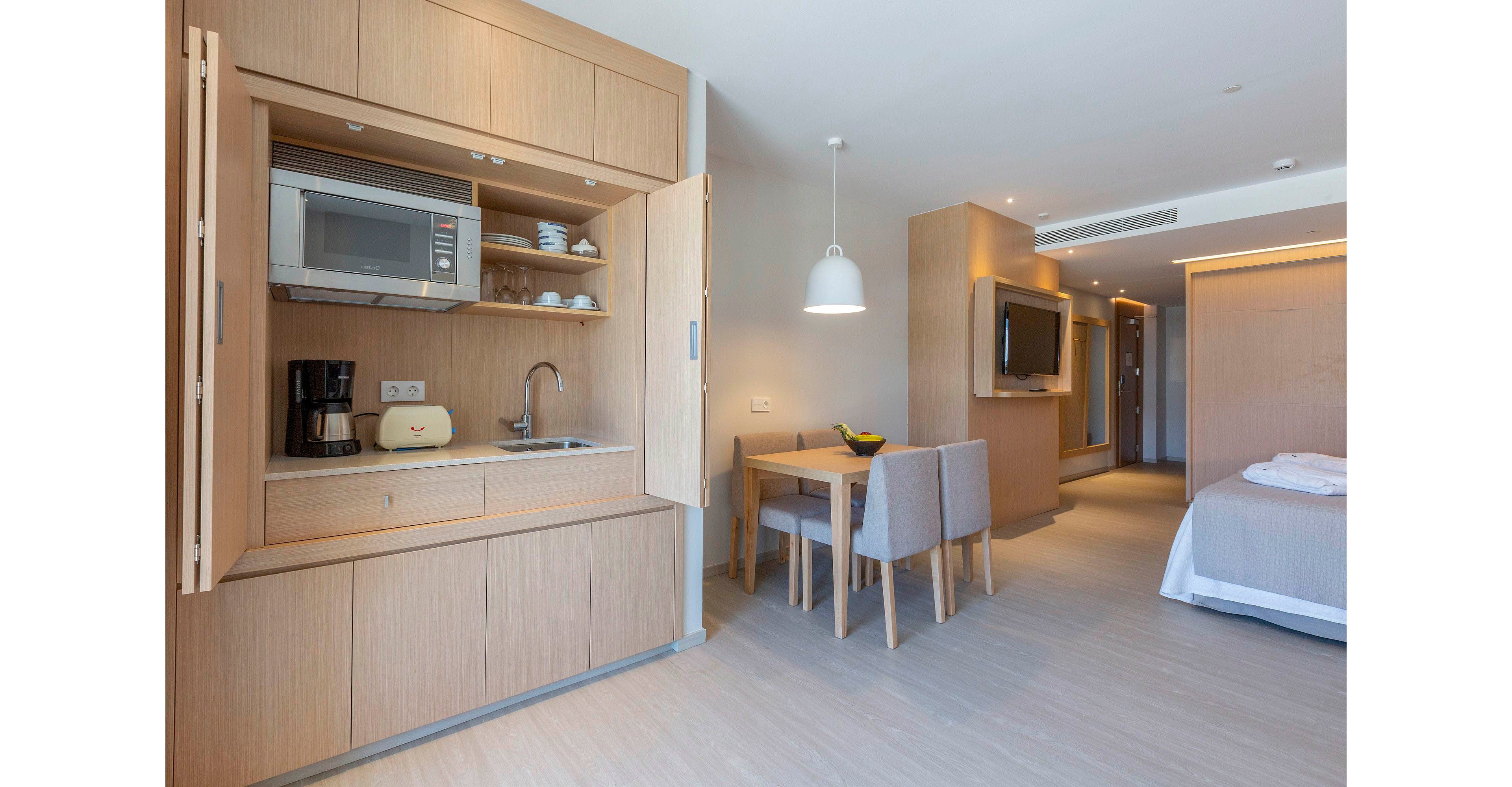 A contemporary hotel suite features a compact kitchenette built into light wood cabinetry. The kitchenette includes a microwave, coffee maker, toaster, sink, and neatly arranged dishes on open shelves. Nearby, a small dining table with upholstered chairs sits under a hanging pendant light. In the background, a wall-mounted television faces the sleeping area with a neatly made bed, creating a clean and minimalist atmosphere.