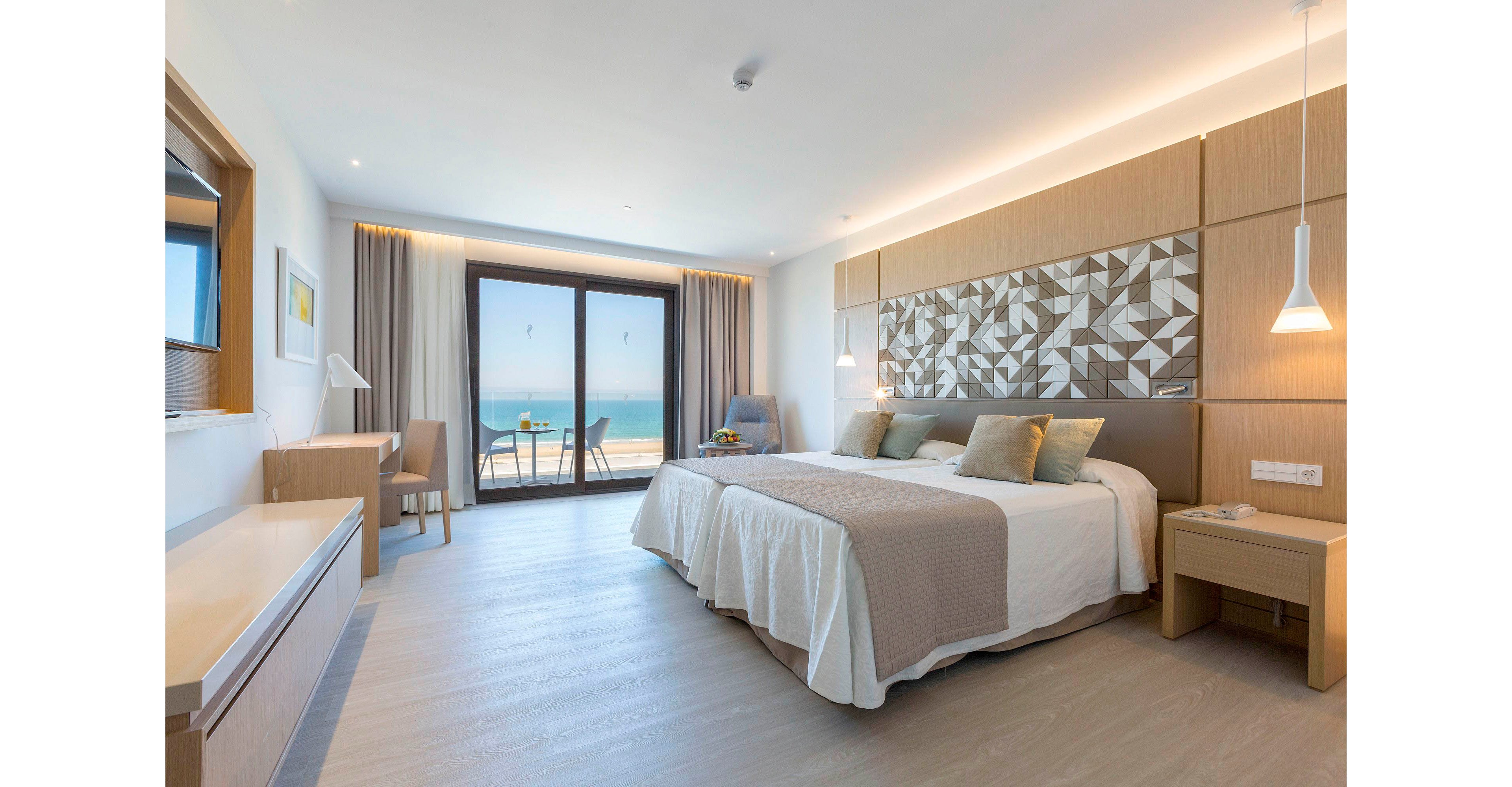 A spacious modern hotel room features a large double bed with neutral-toned bedding and decorative geometric wall paneling. Floor-to-ceiling glass doors open onto a balcony overlooking a bright blue ocean. Light wood furniture, including bedside tables, a desk, and a dresser, complement the soft beige and white color palette. Pendant lights and recessed ceiling lighting create a calm, inviting atmosphere. A small table and chairs on the balcony suggest a relaxing spot to enjoy the coastal view.