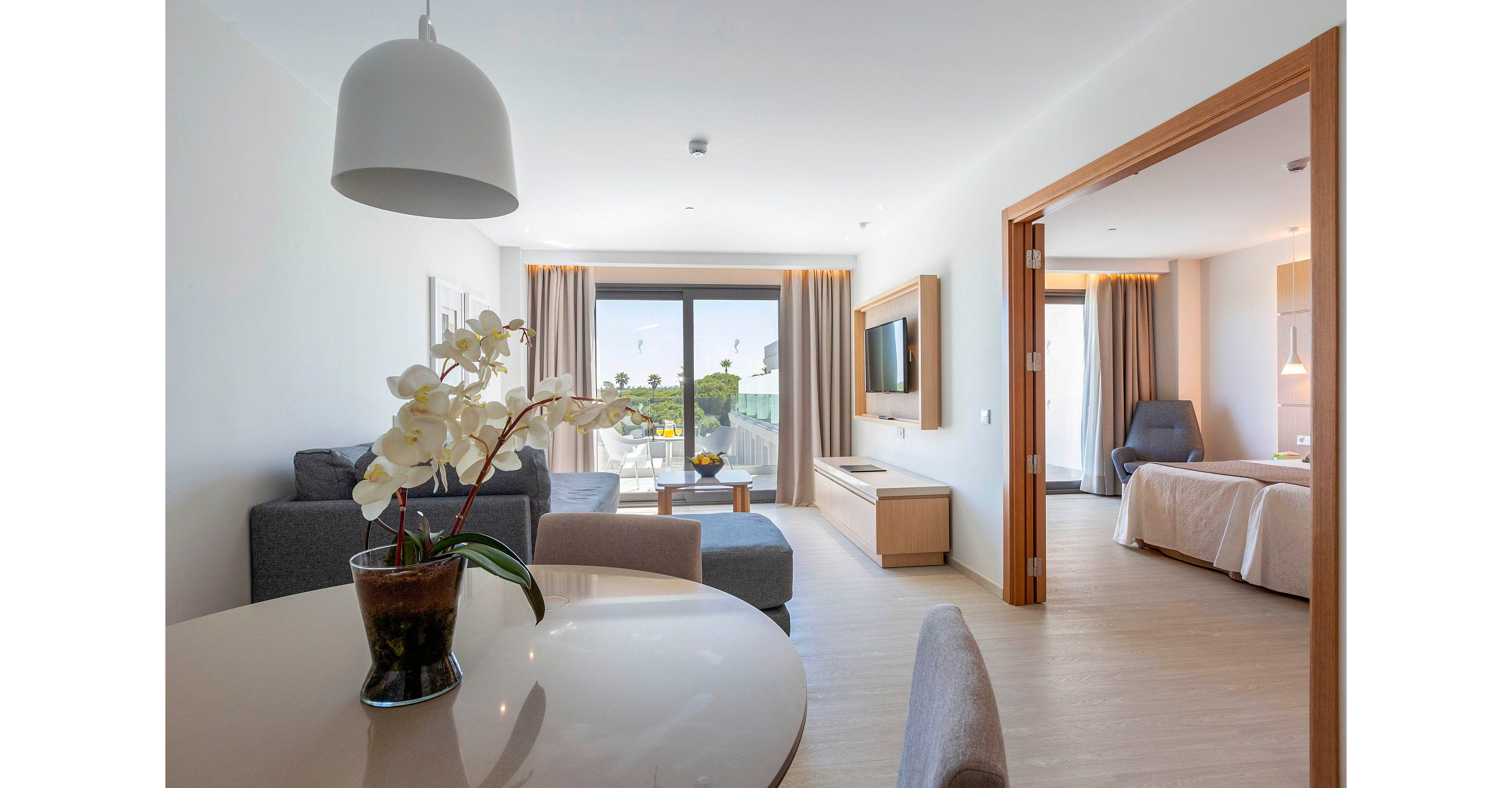 A contemporary hotel suite features an open-plan living and dining area with neutral-toned furniture and a large glass door leading to a balcony. A round dining table with a vase of white orchids sits in the foreground, while a gray sofa and ottoman face a wall-mounted television. Beyond the sliding doors, an outdoor seating area overlooks a sunny landscape with palm trees. The bedroom area is partially visible through a wide wooden-framed opening, showing a neatly made bed and an armchair by the window. Soft natural light and minimalist decor create a calm, inviting atmosphere.