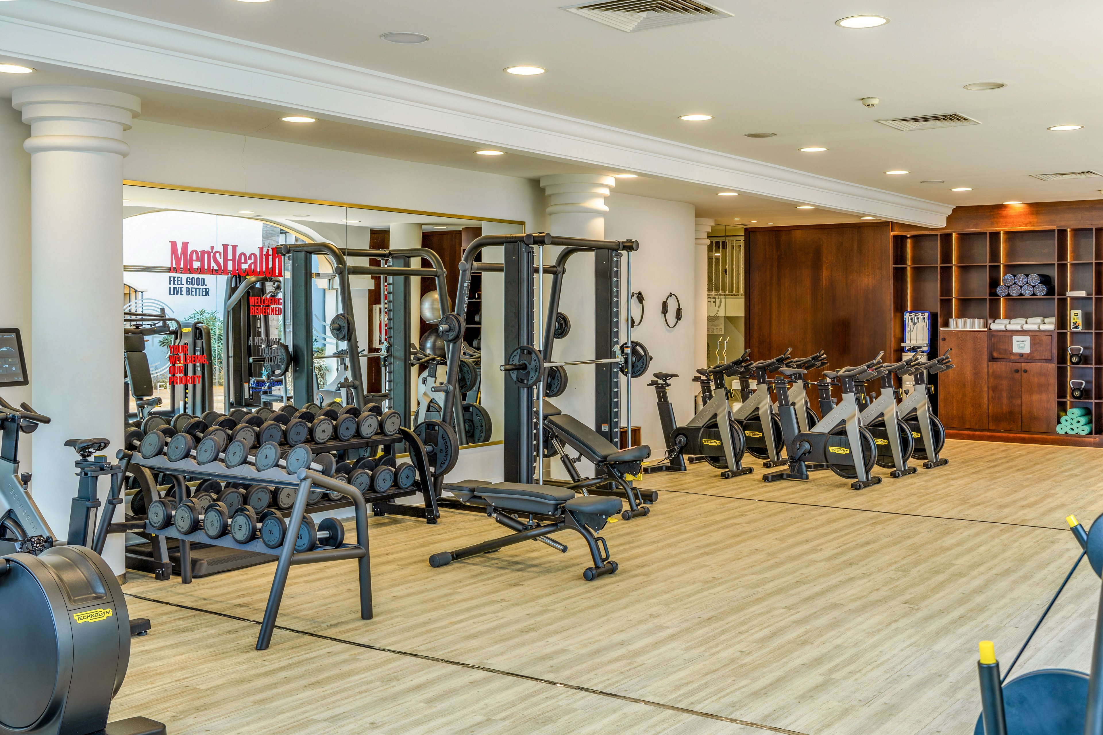 Spacious indoor gym featuring free weights, benches, and strength machines. The room has light wood flooring, white columns, and large mirrors along the wall. Cardio equipment is visible in the foreground, with neatly organized dumbbells and weight racks in the center. A wooden shelving unit and subtle decor create a clean, contemporary workout environment.