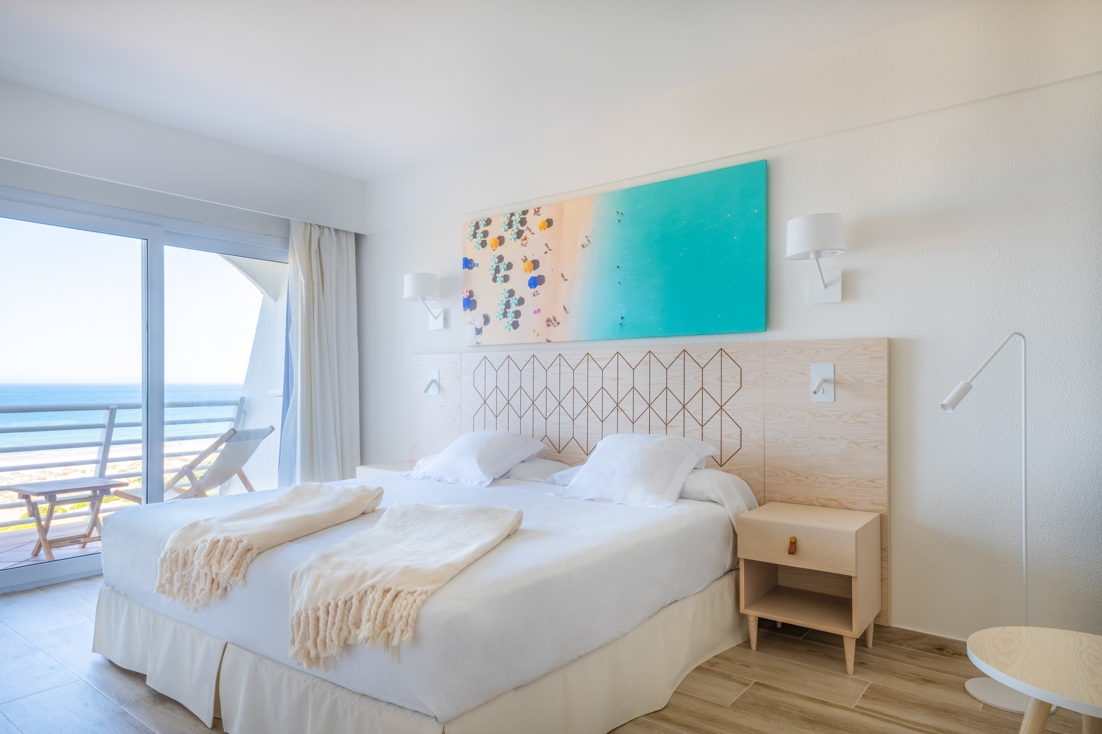 A modern hotel room featuring a large bed with white linens and beige throws, complemented by light wood furniture. The room has a balcony with a stunning ocean view, visible through a glass sliding door. The decor includes a colorful abstract painting above the bed, adding vibrancy to the serene setting.