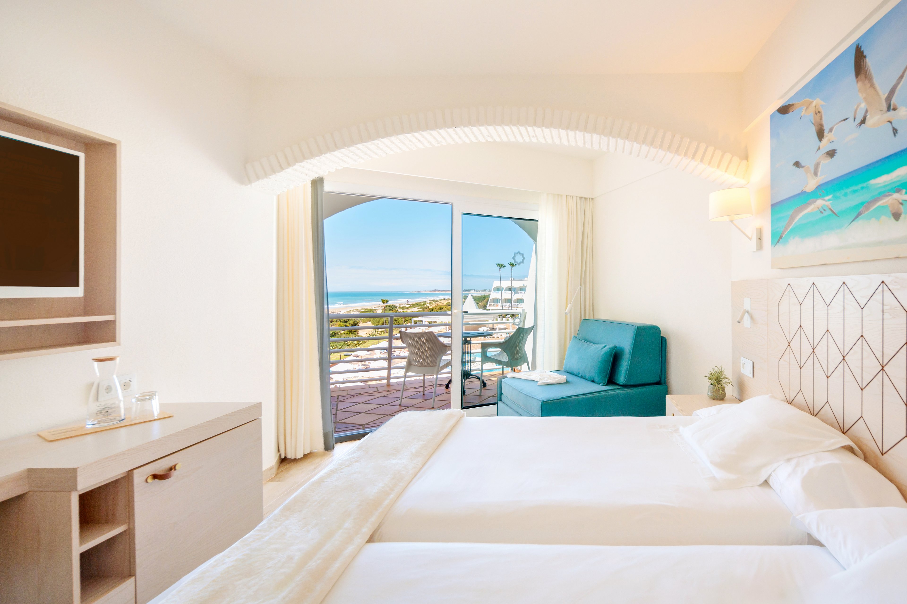 A modern hotel room featuring two neatly made beds, a teal armchair, and a wall-mounted TV. The room opens to a balcony with a stunning ocean view, showcasing clear skies and a serene landscape. Decor includes a vibrant seagull painting and geometric wall patterns, creating a relaxing atmosphere.