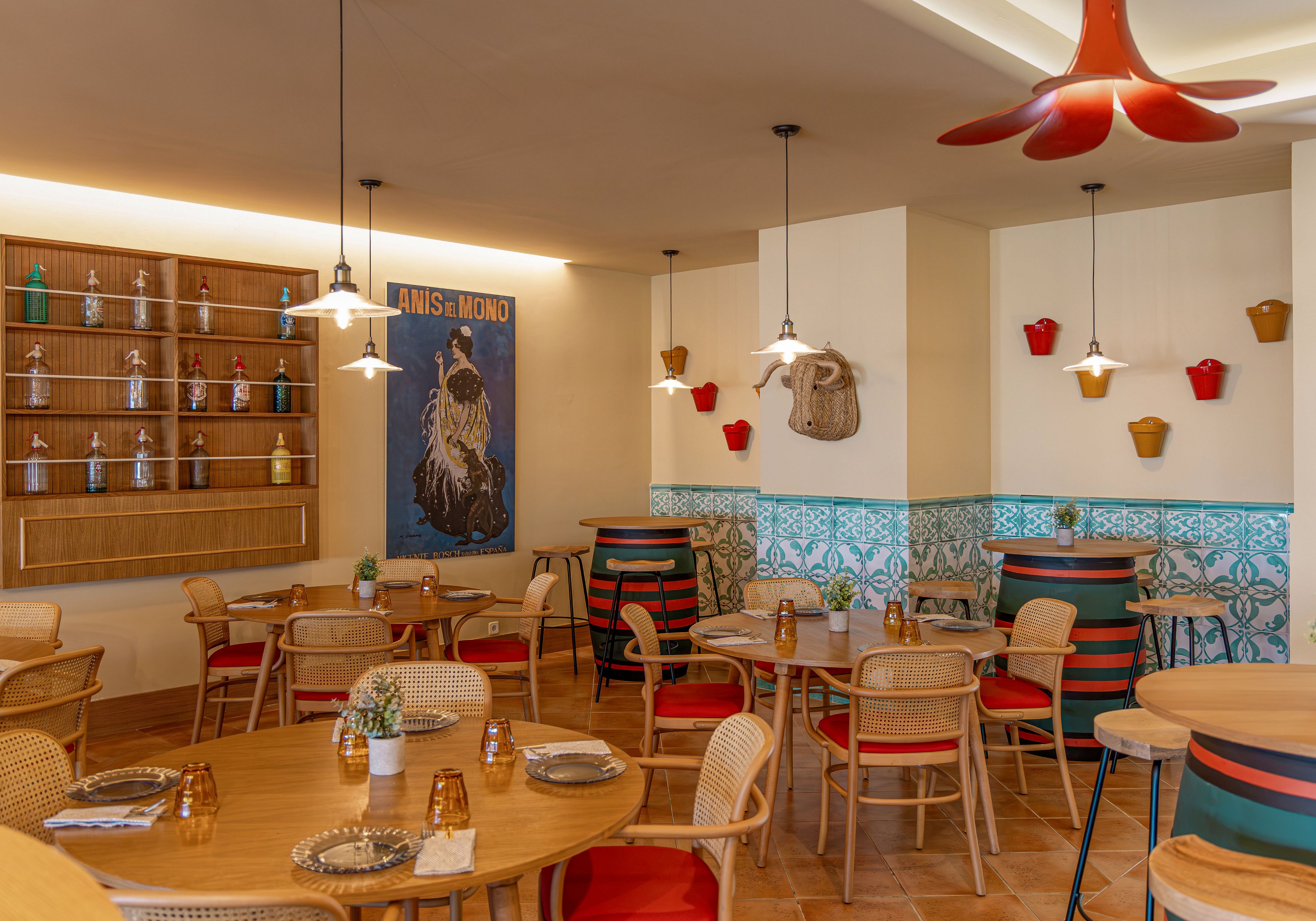 Warmly lit café interior featuring wooden tables and wicker chairs arranged for casual dining. Colorful barrel-style tables and patterned turquoise wall tiles add a playful Mediterranean touch. Decorative shelves with bottles, hanging planters, and unique pendant lights create visual interest. A large wall poster reading "ANIS MONO" serves as a focal point in the room.