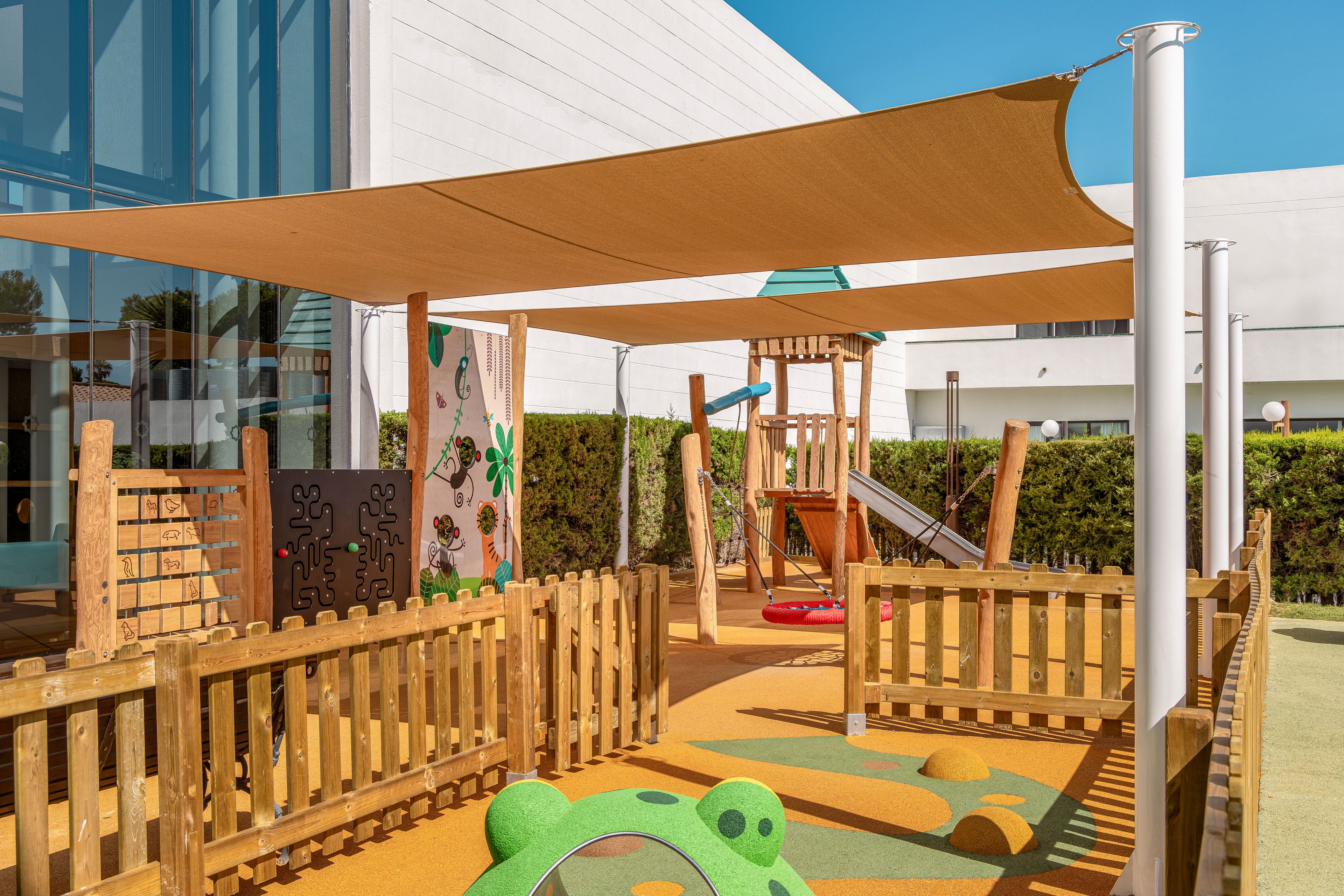 Contemporary outdoor playground with wooden structures is set beside a modern building with large glass windows. The play area includes climbing equipment, a slide, and soft rubber flooring with colorful shapes. A fabric shade canopy stretches above the fenced space, providing sun protection. Green hedges and clear blue sky frame the bright, clean recreational environment.