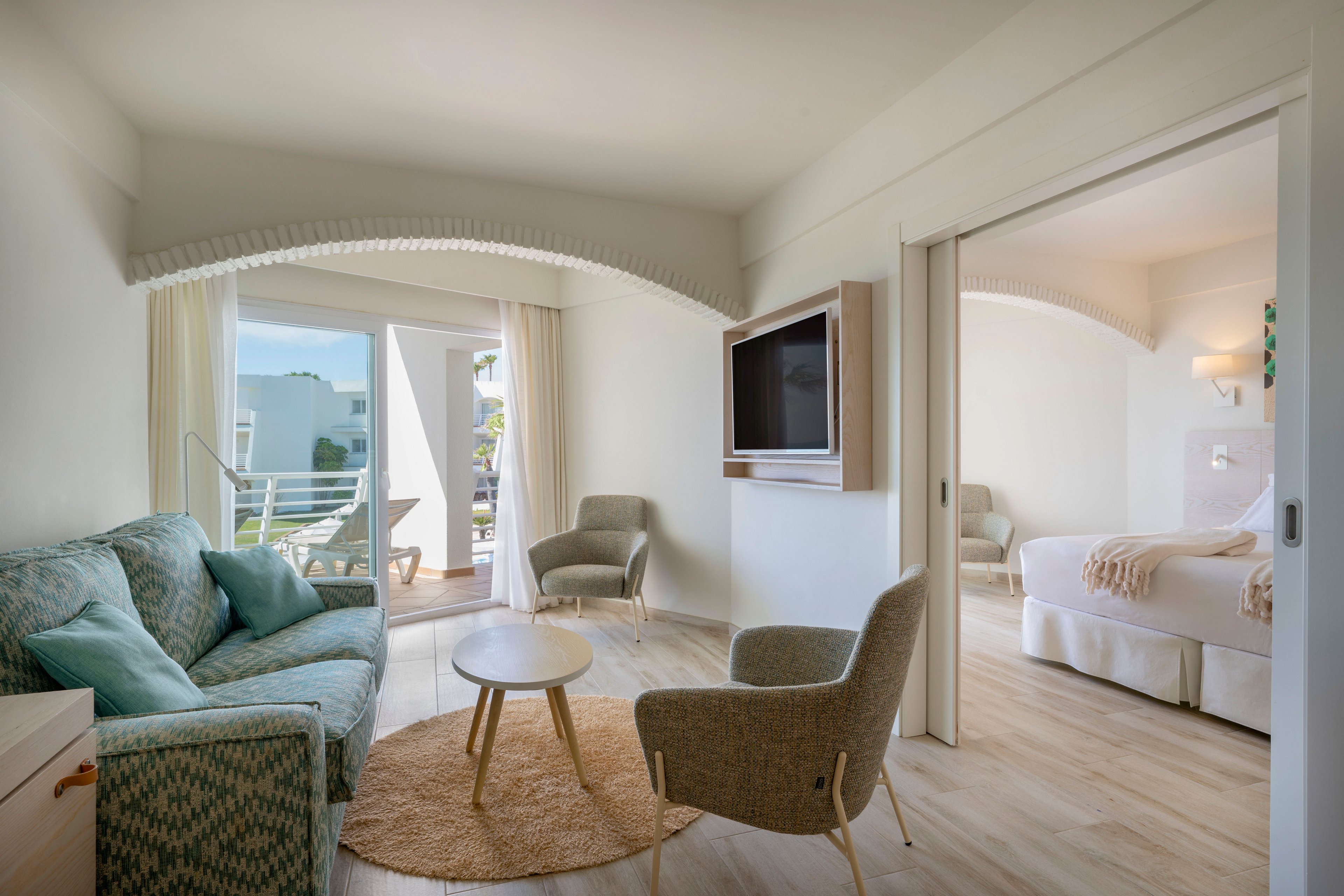 A modern hotel suite features a cozy sitting area with armchairs, a sofa, and a round coffee table on a woven rug. Large sliding doors open to a balcony, allowing natural light to fill the room and reveal an outdoor view. A wall-mounted flat-screen television faces the seating area, while a separate bedroom with a double bed is visible through a wide doorway. The interior design is airy and minimalist, with light wood flooring and soft neutral tones creating a relaxed, coastal atmosphere.