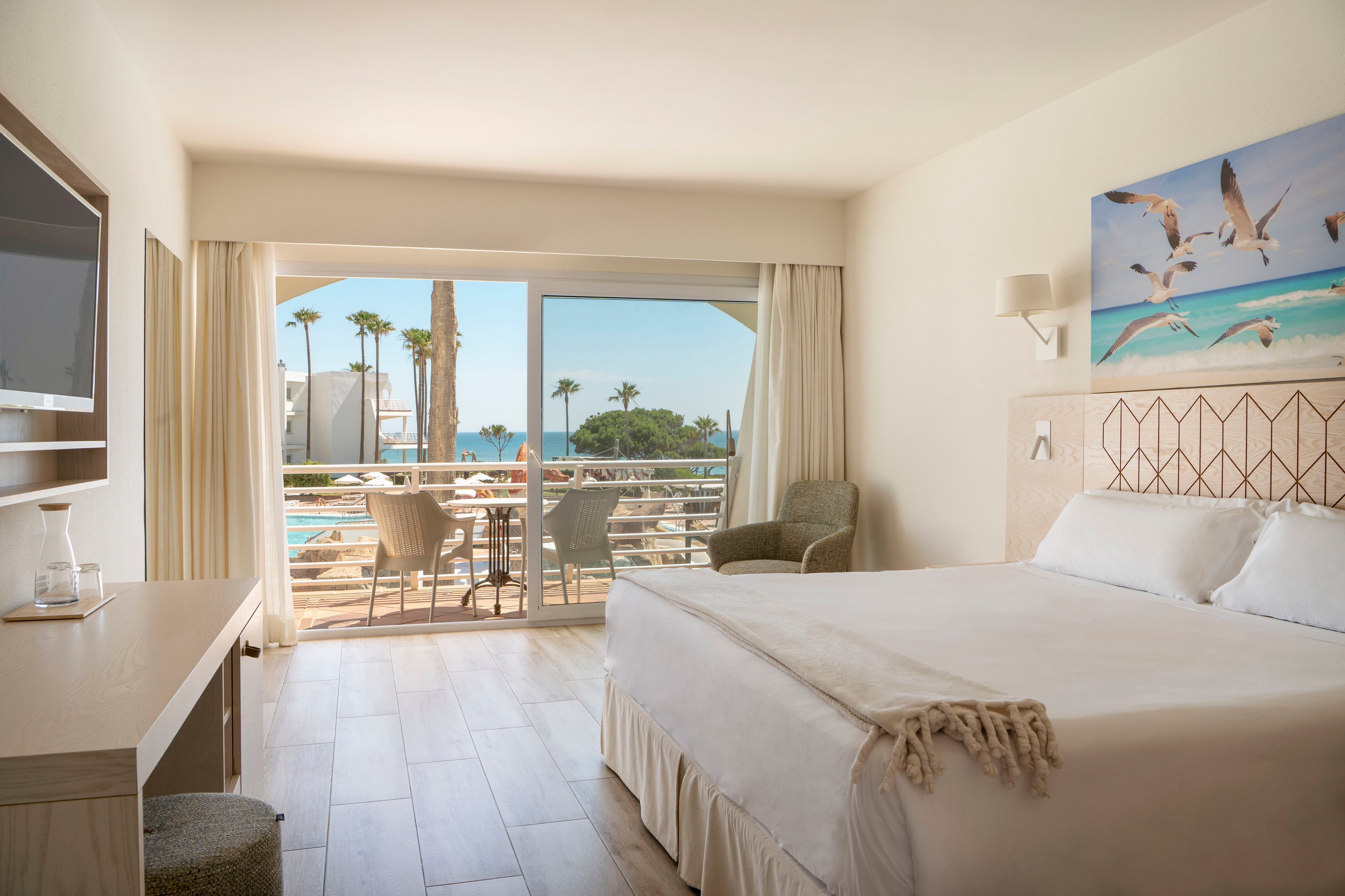 A sunlit hotel room features a neatly made double bed facing a balcony with sliding glass doors. Outside, a terrace with chairs overlooks a pool area, palm trees, and the ocean in the distance. The interior decor is light and neutral, with wooden flooring, a desk, and an armchair creating a relaxed atmosphere. A large wall art of flying seagulls above the bed reinforces the seaside vacation setting.