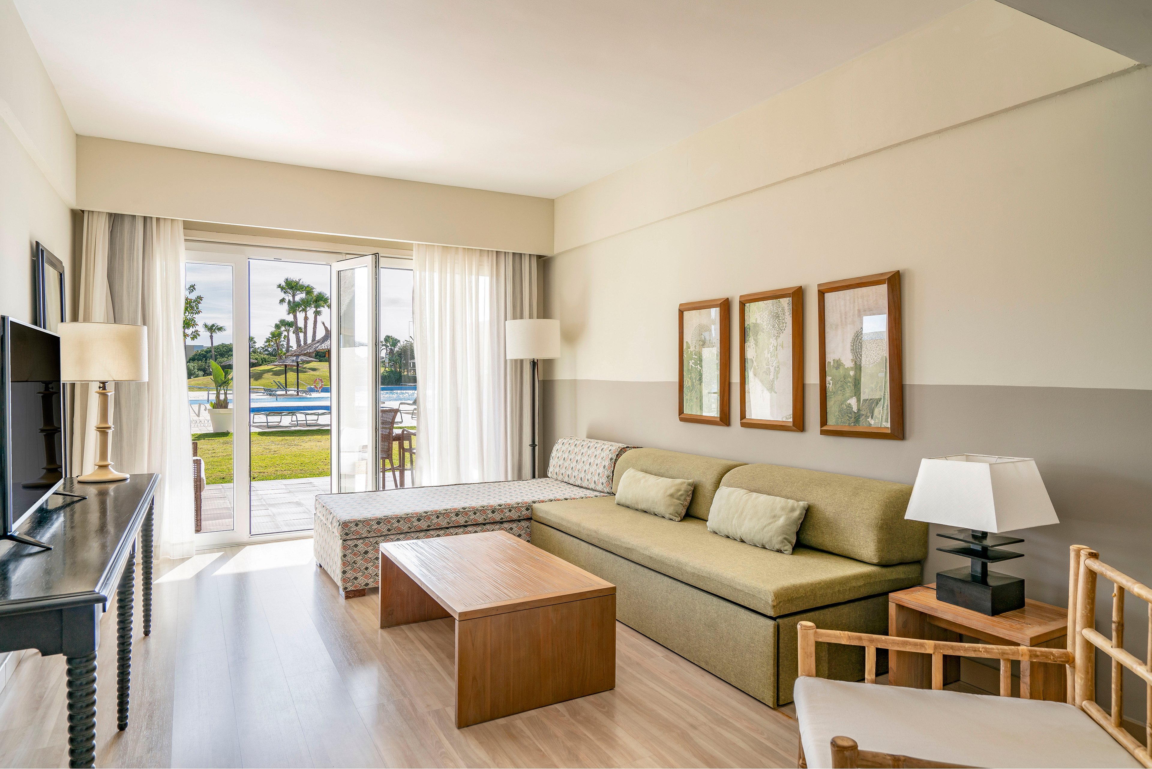 A modern hotel suite features a long green sofa with cushions and a wooden coffee table in the center. Large glass doors open to a terrace overlooking a pool and palm trees, allowing natural light to fill the room. Neutral tones, light wood flooring, and minimalist framed artwork create a calm, contemporary atmosphere. A television, lamps, and simple chairs complete the comfortable vacation setting.