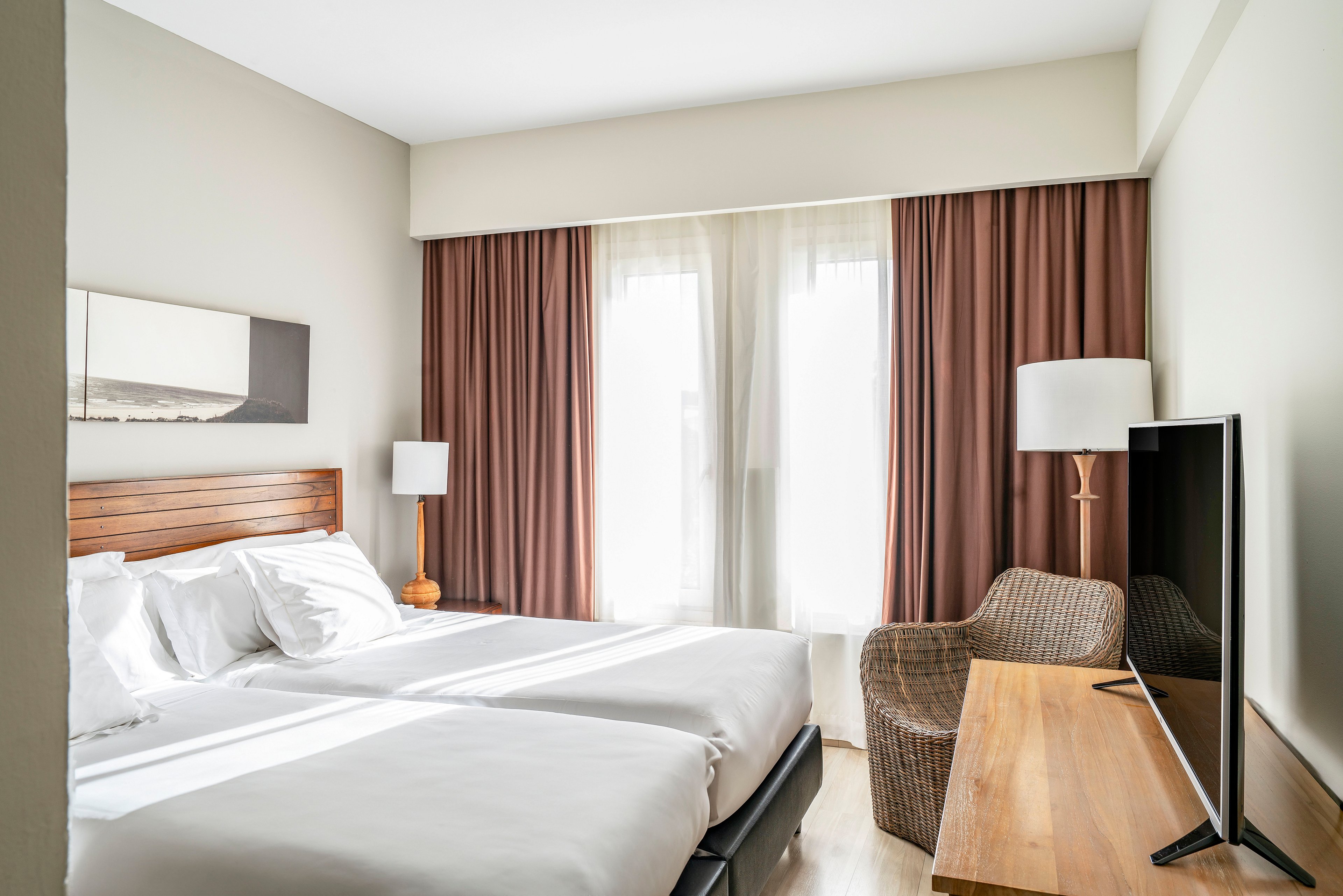 A bright, modern hotel room features two neatly made twin beds with white linens and a wooden headboard. Large windows with sheer curtains and brown drapes allow natural light to fill the space. A wicker armchair and floor lamp sit beside a wooden desk that holds a flat-screen television. The neutral color palette and simple decor create a calm, minimalist atmosphere suitable for relaxation or business travel.