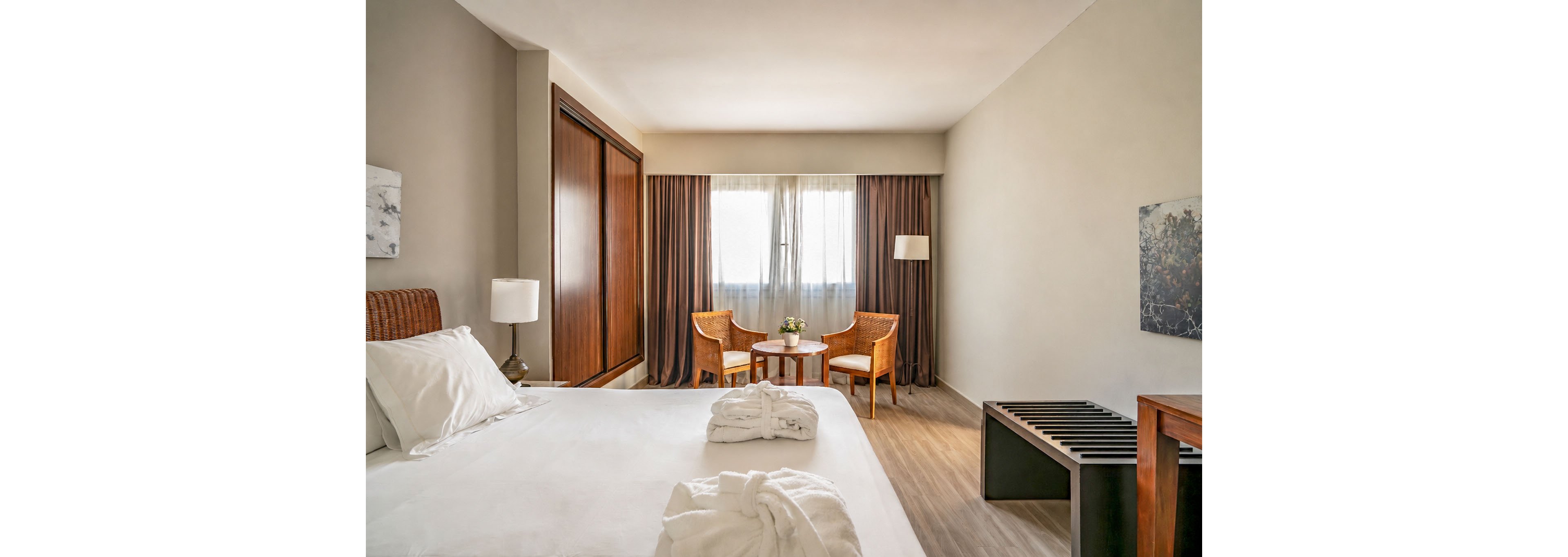 A neatly made bed with white linens and folded bathrobes is the focal point of a modern hotel room. The space features a small seating area with two woven chairs and a round table near a large window with sheer and blackout curtains. Warm wooden furniture, including a wardrobe, bench, and bedside tables, contrasts with the neutral walls and flooring. Soft lighting and minimal decor create a clean, inviting atmosphere suitable for relaxation or business travel.