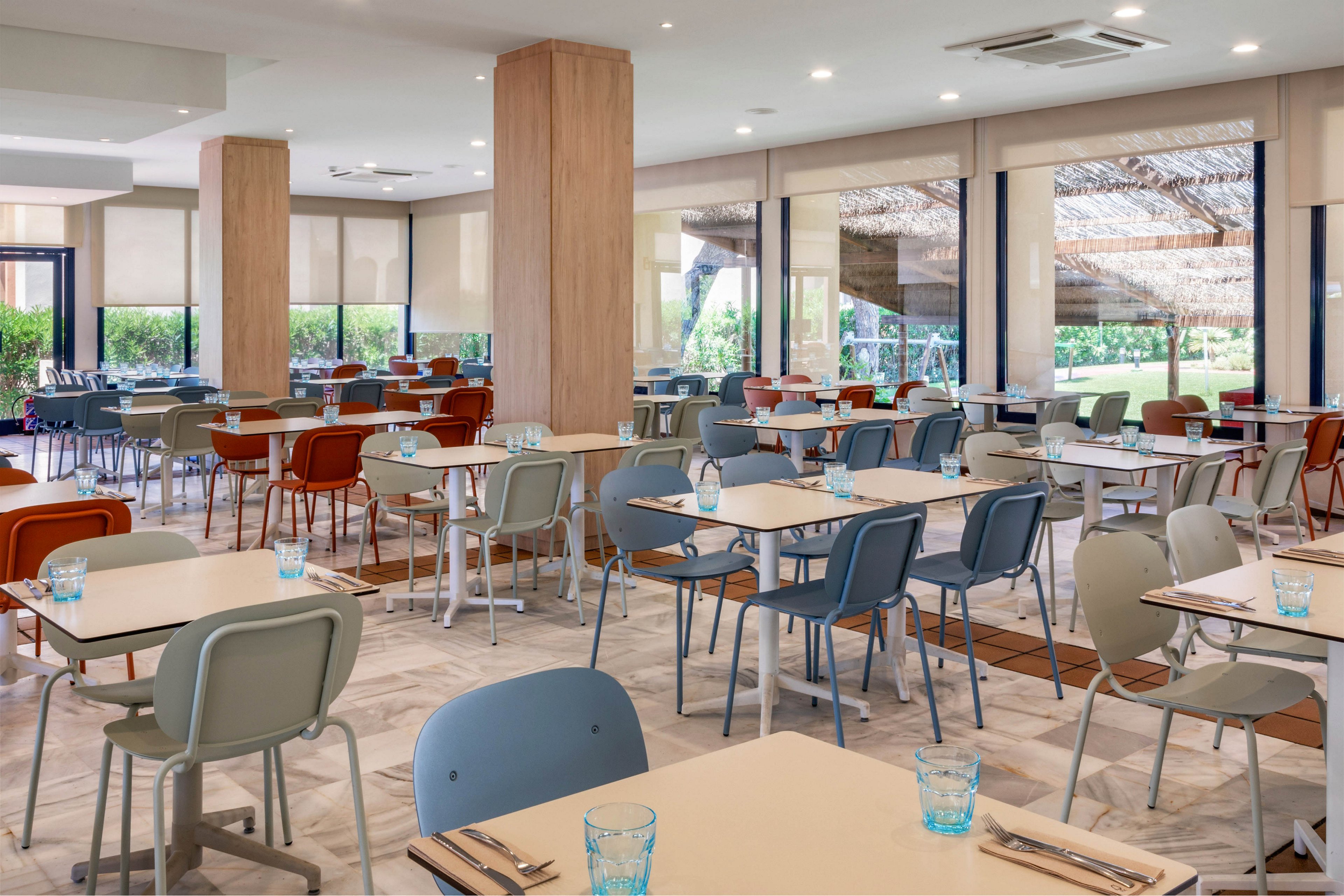 A contemporary restaurant dining room is shown empty and neatly arranged. Light-colored tables are paired with blue, gray, and brown chairs, each set with cutlery and blue glass tumblers. Large windows with roller shades let in natural light and offer views of greenery outside. Neutral tones, clean lines, and ample space create a calm, inviting atmosphere suitable for casual or hotel dining.