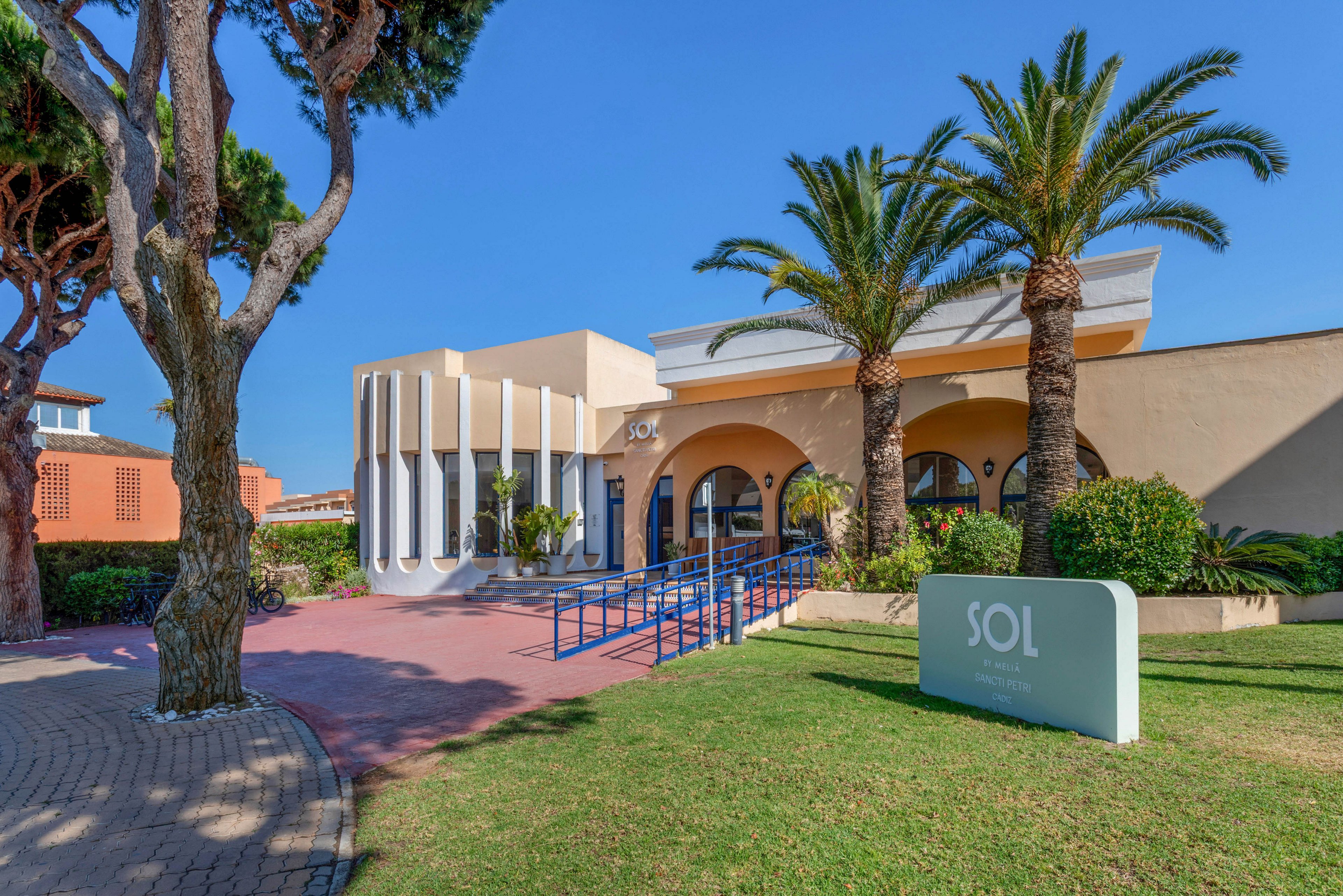A contemporary low-rise building with arched entrances and geometric columns stands under a clear blue sky. The structure features a light beige and white facade, accented by blue railings and a red-tiled walkway. Tall palm trees and manicured shrubs frame the entrance, creating a relaxed Mediterranean atmosphere. A freestanding sign in the foreground displays the word SOL, indicating the name of the property or venue.